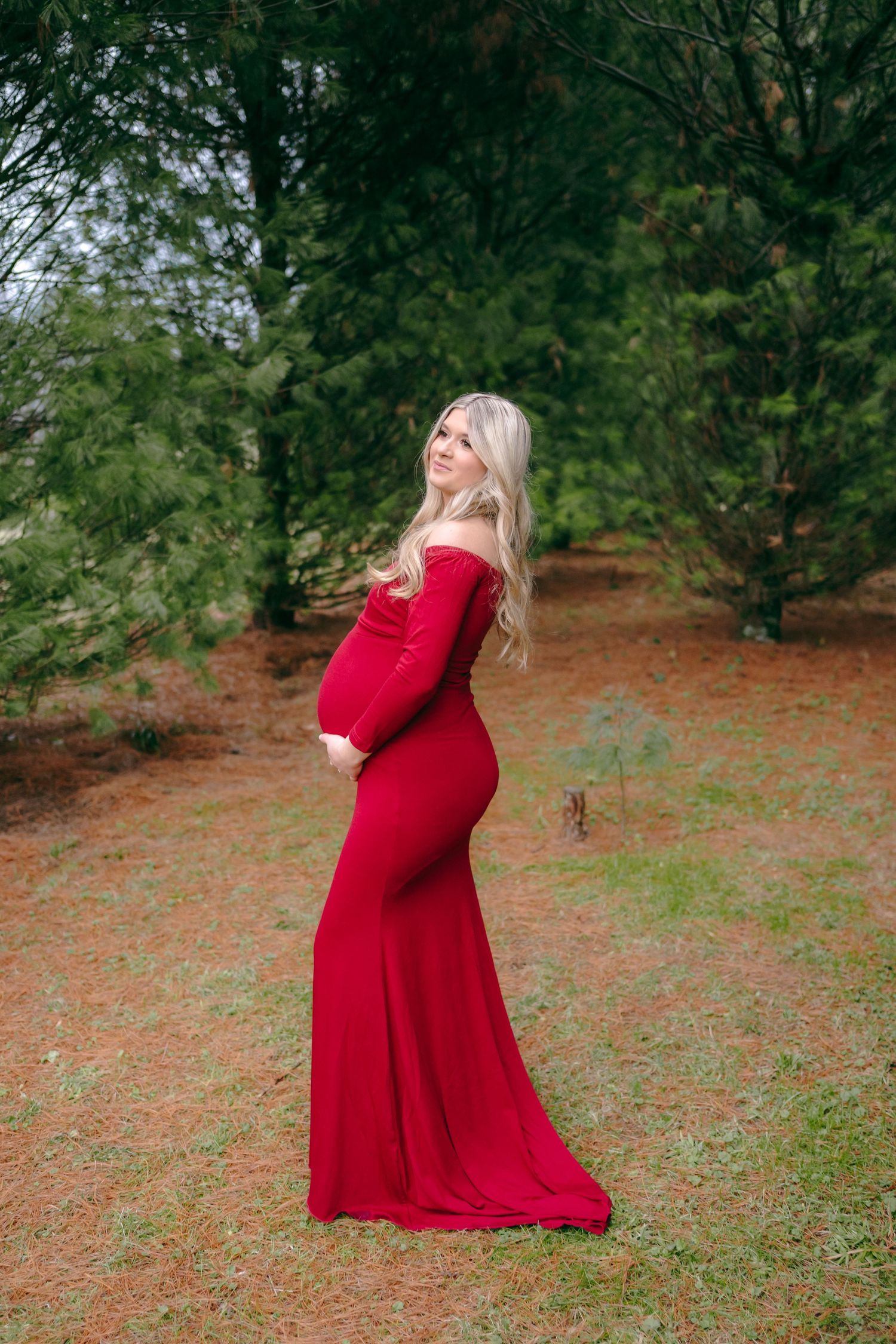 Woman in flowing red dress shows off baby bump at Christmas tree farm.
