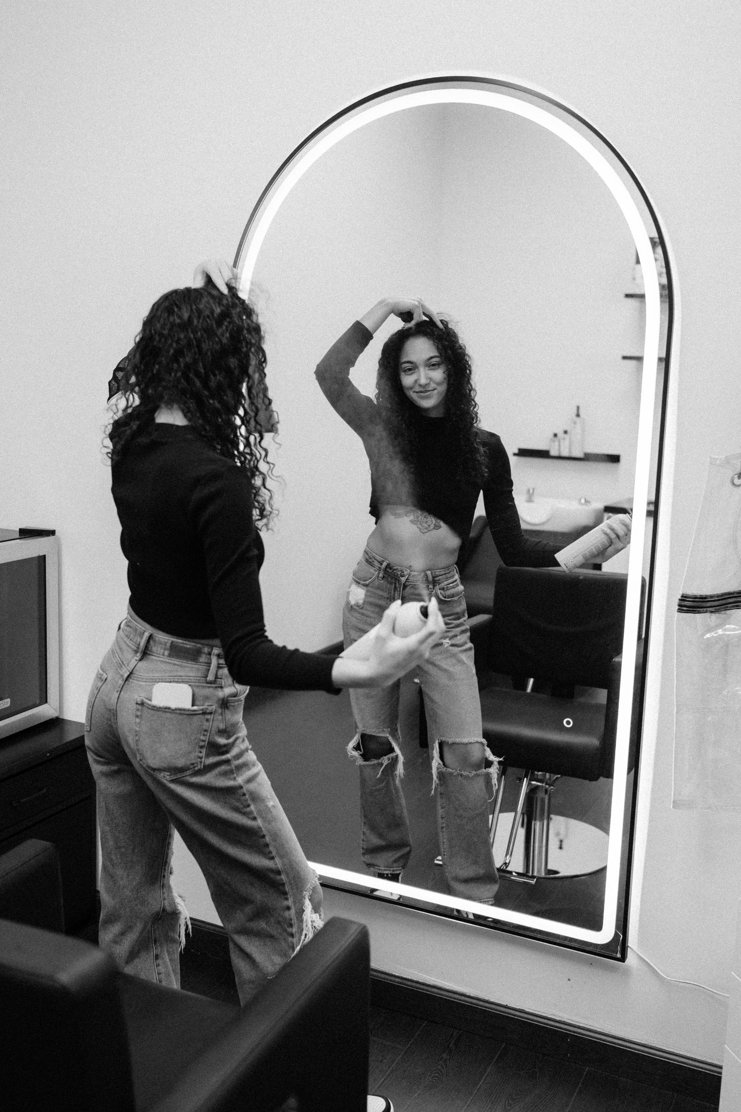 Person posing playfully in front of an arched mirror wearing jeans and a black top.