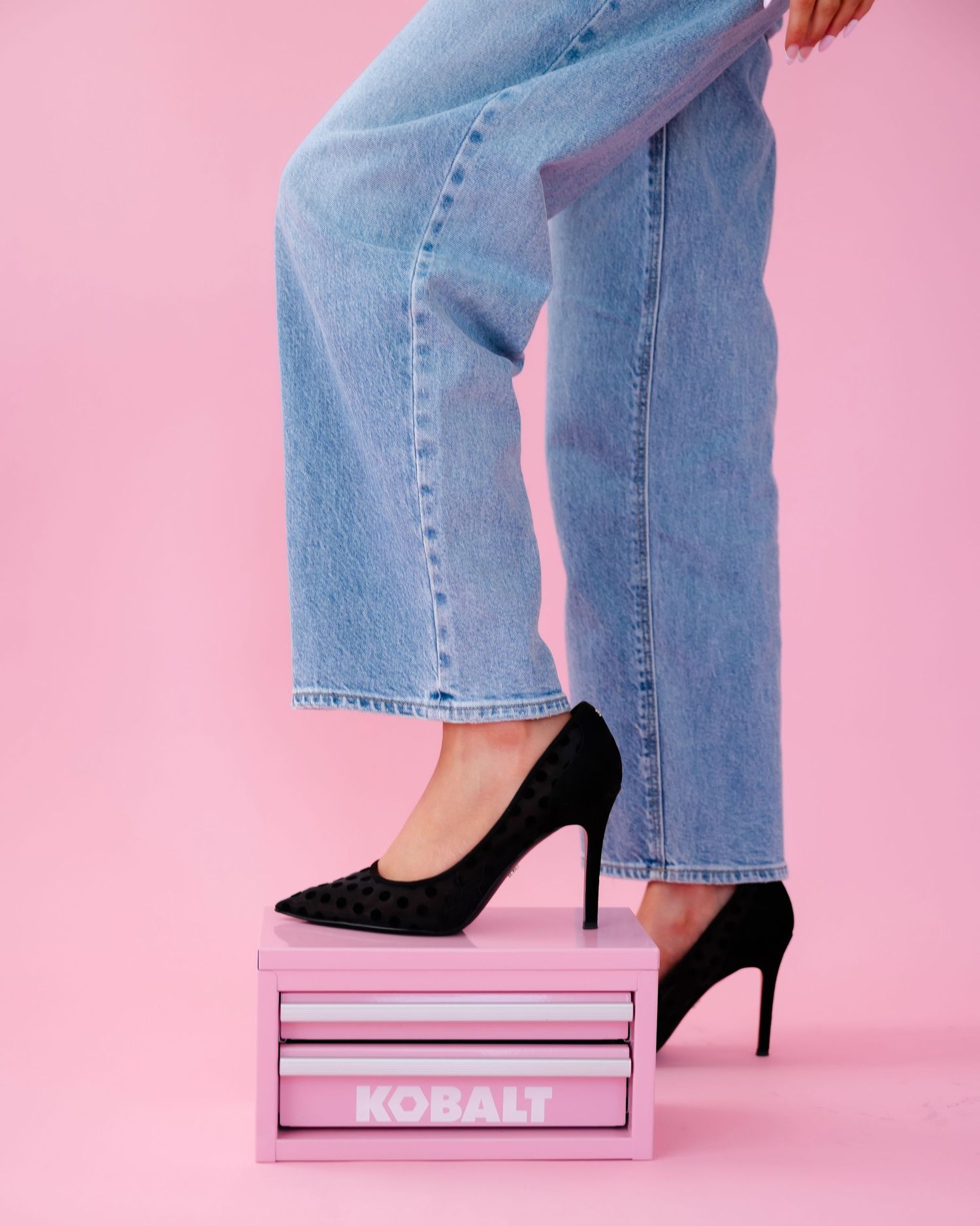 Close-up of denim jeans and black stiletto heels against pink backdrop showing modern fashion styling.