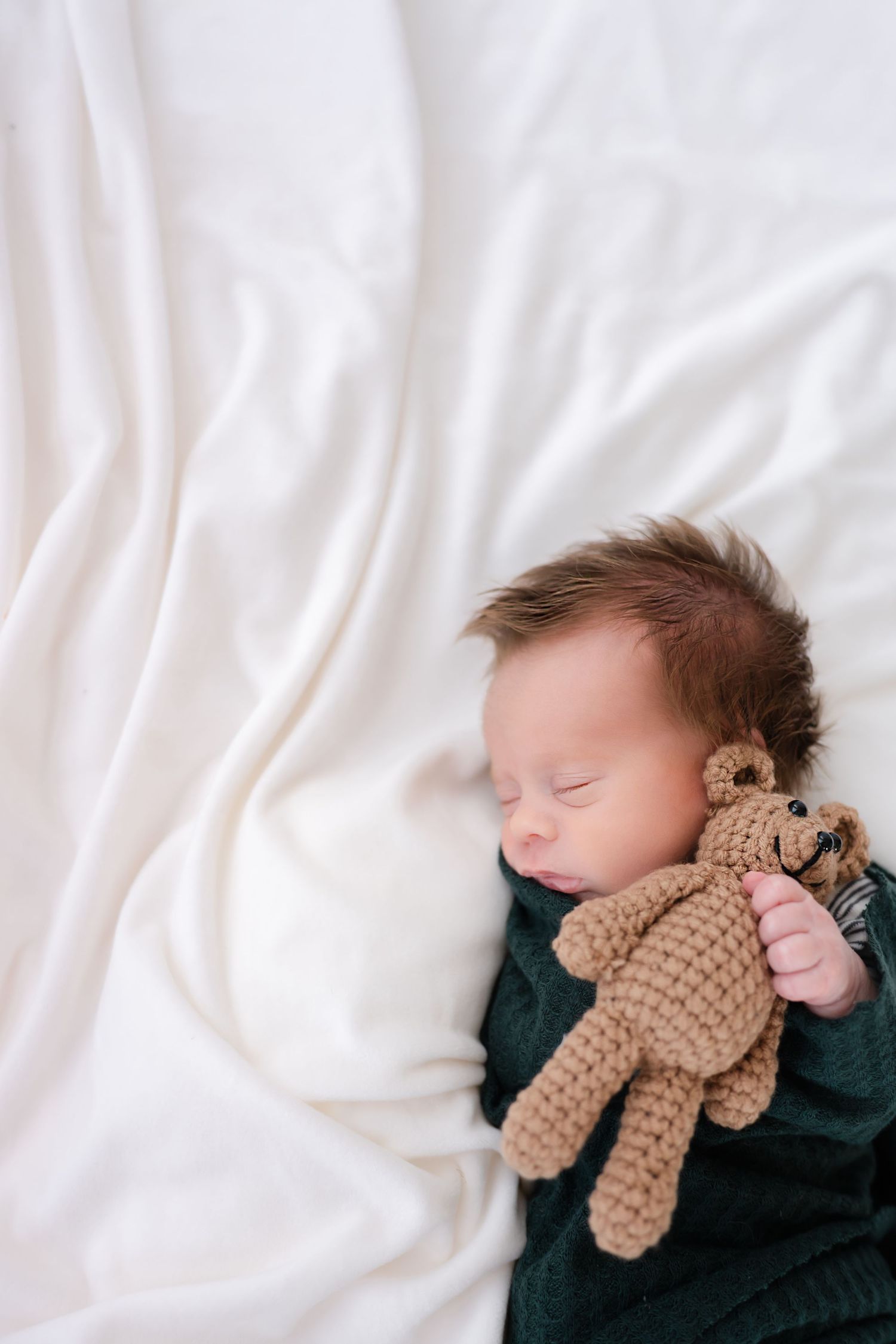 A sleeping baby in dark clothing cuddles with a small brown teddy bear on white bedding.