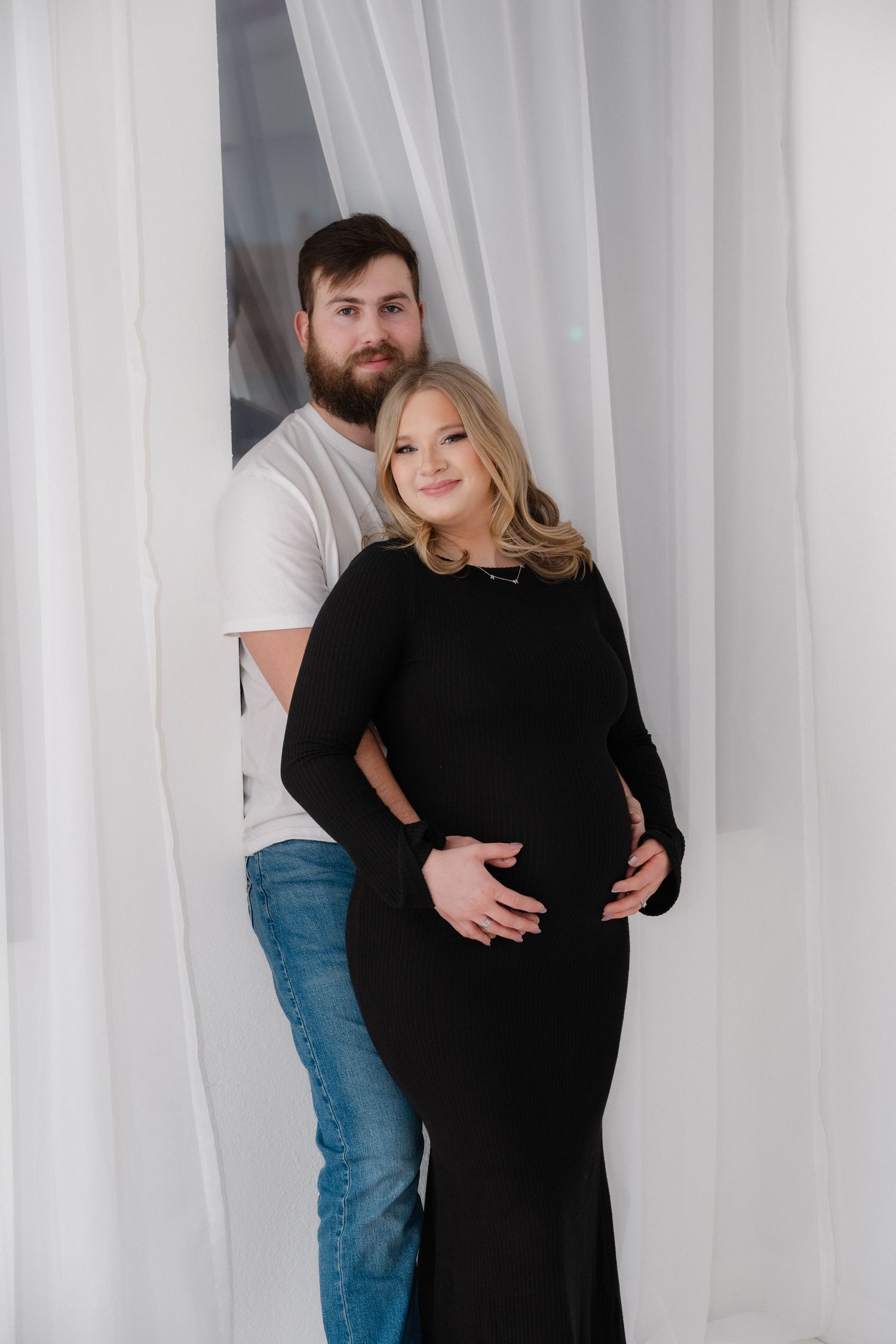Couple embracing during maternity photoshoot with white curtains in background.