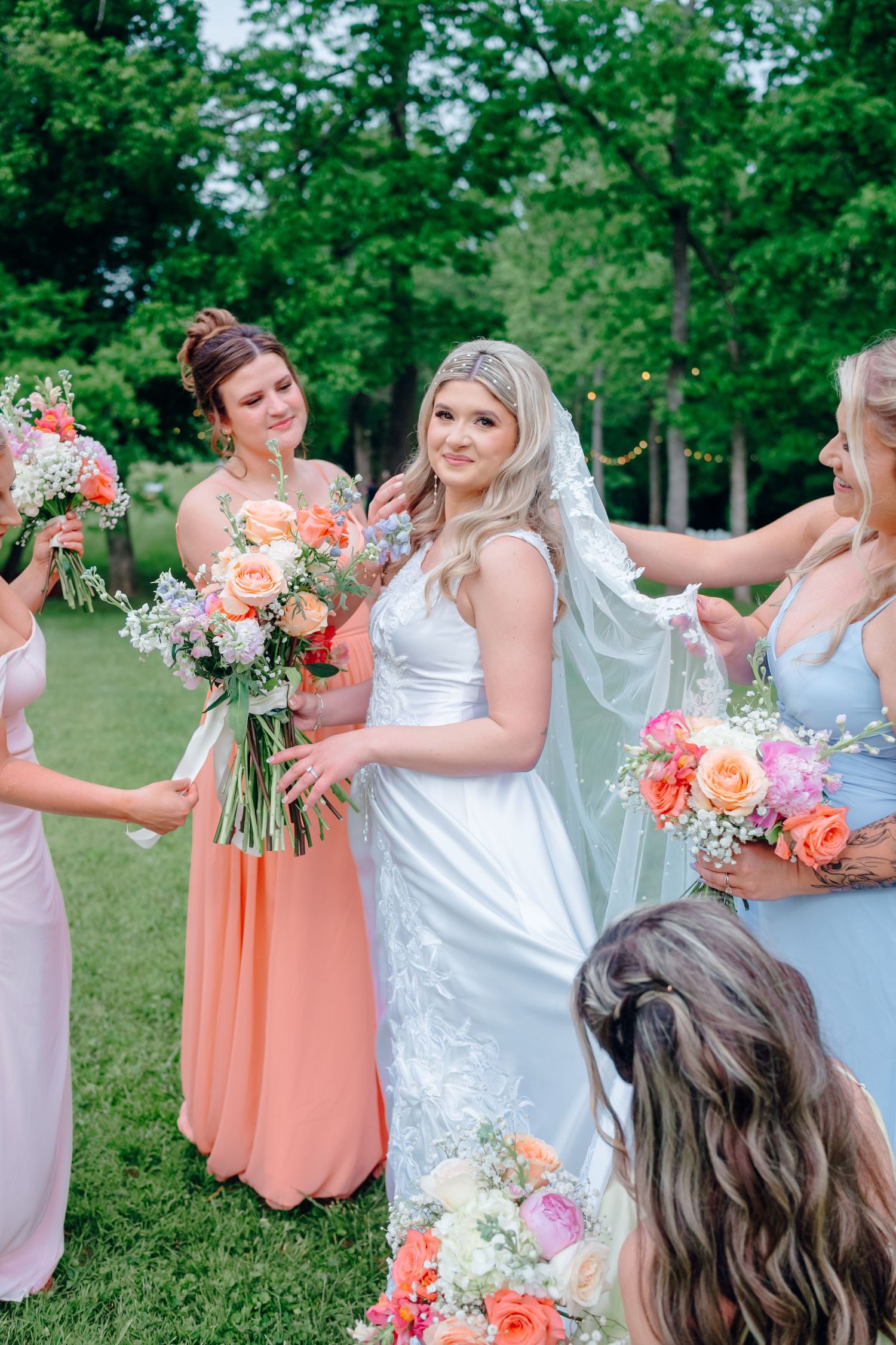 Bridesmaids in pastel dresses holding coral and pink bouquets outdoors.