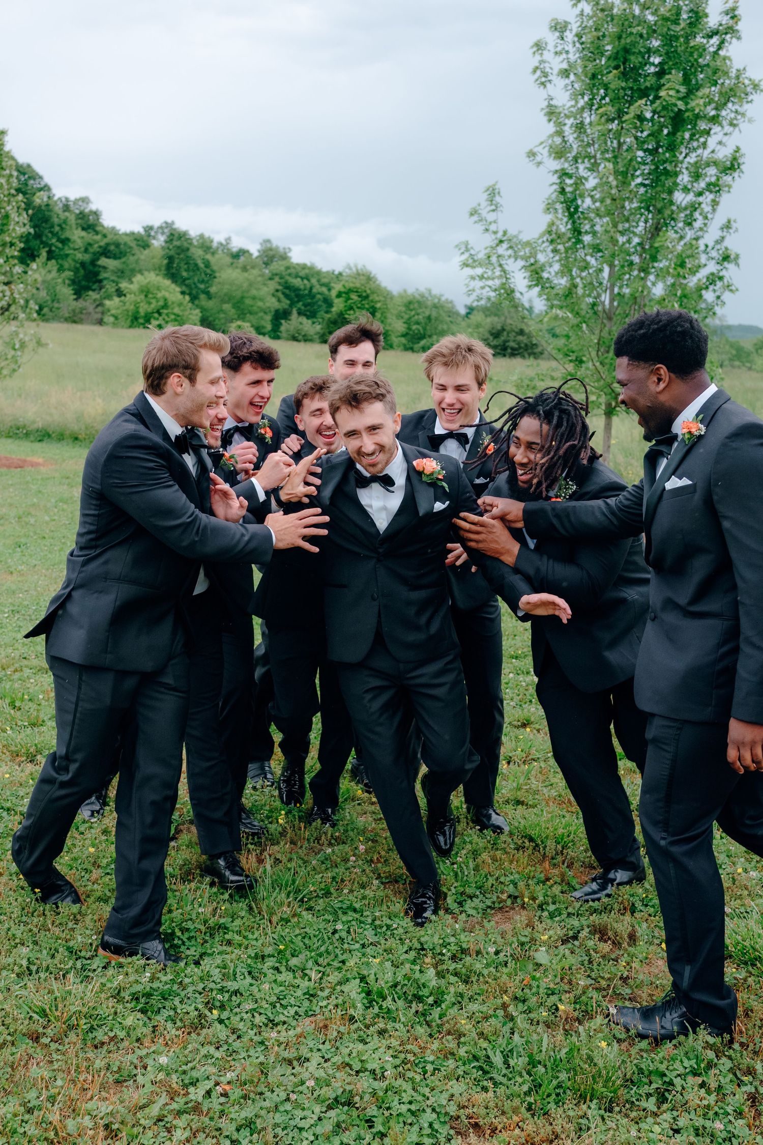 Groomsmen in black suits pose playfully in a line on grassy field during wedding photoshoot.
