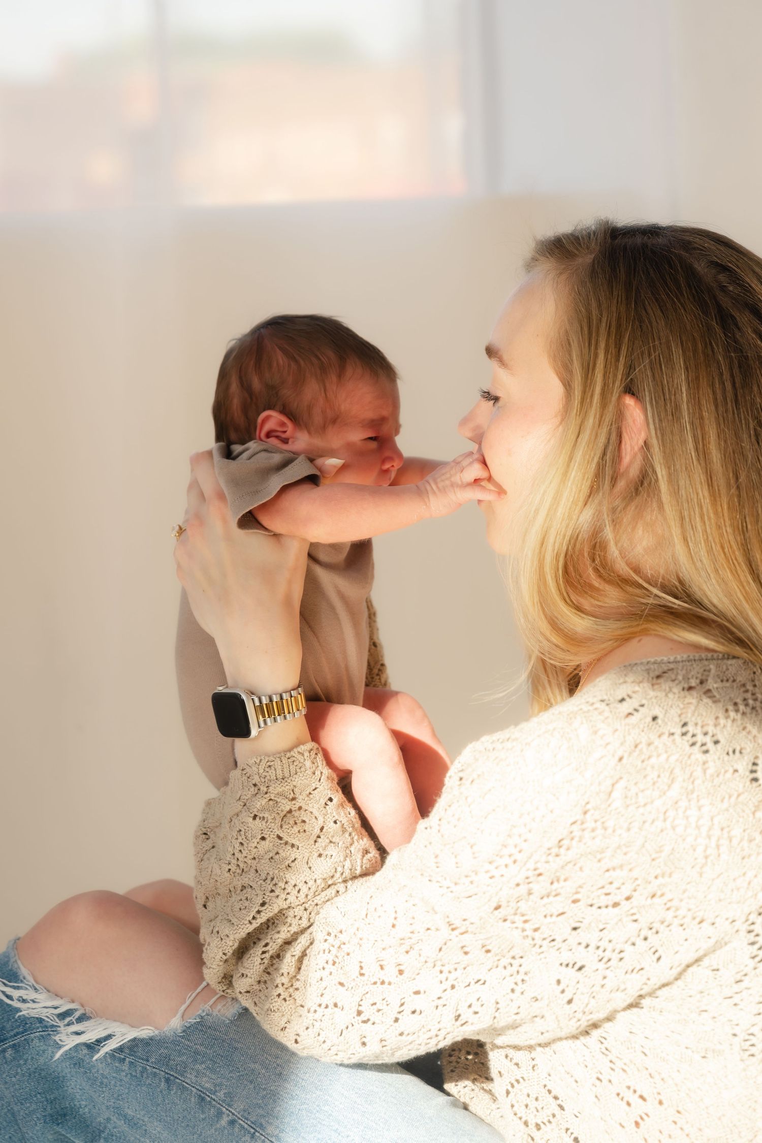 A young blonde mother lovingly holds her newborn baby up to her face in warm natural light.
