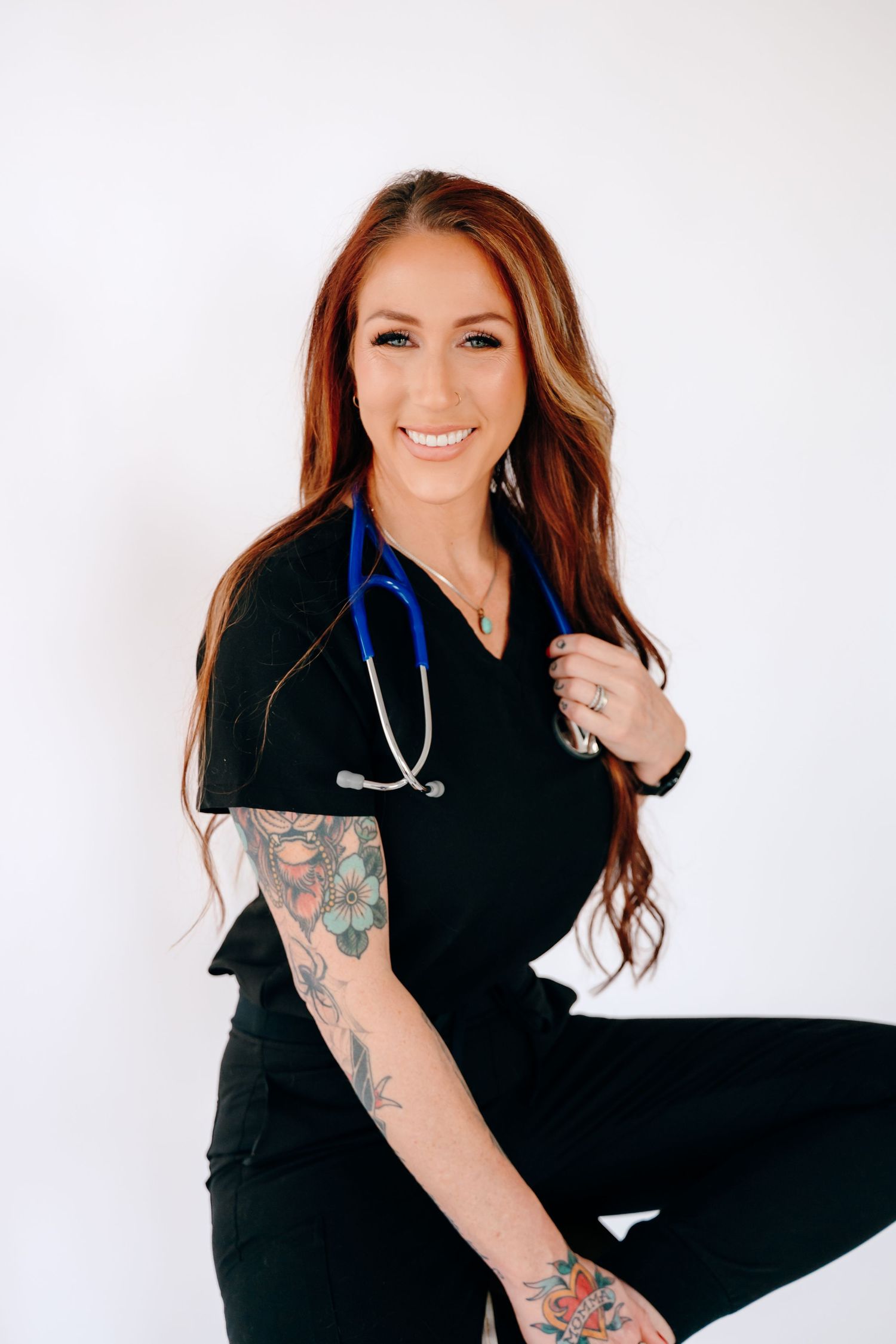 Healthcare professional in black scrubs with colorful arm tattoos and a stethoscope poses against white background.