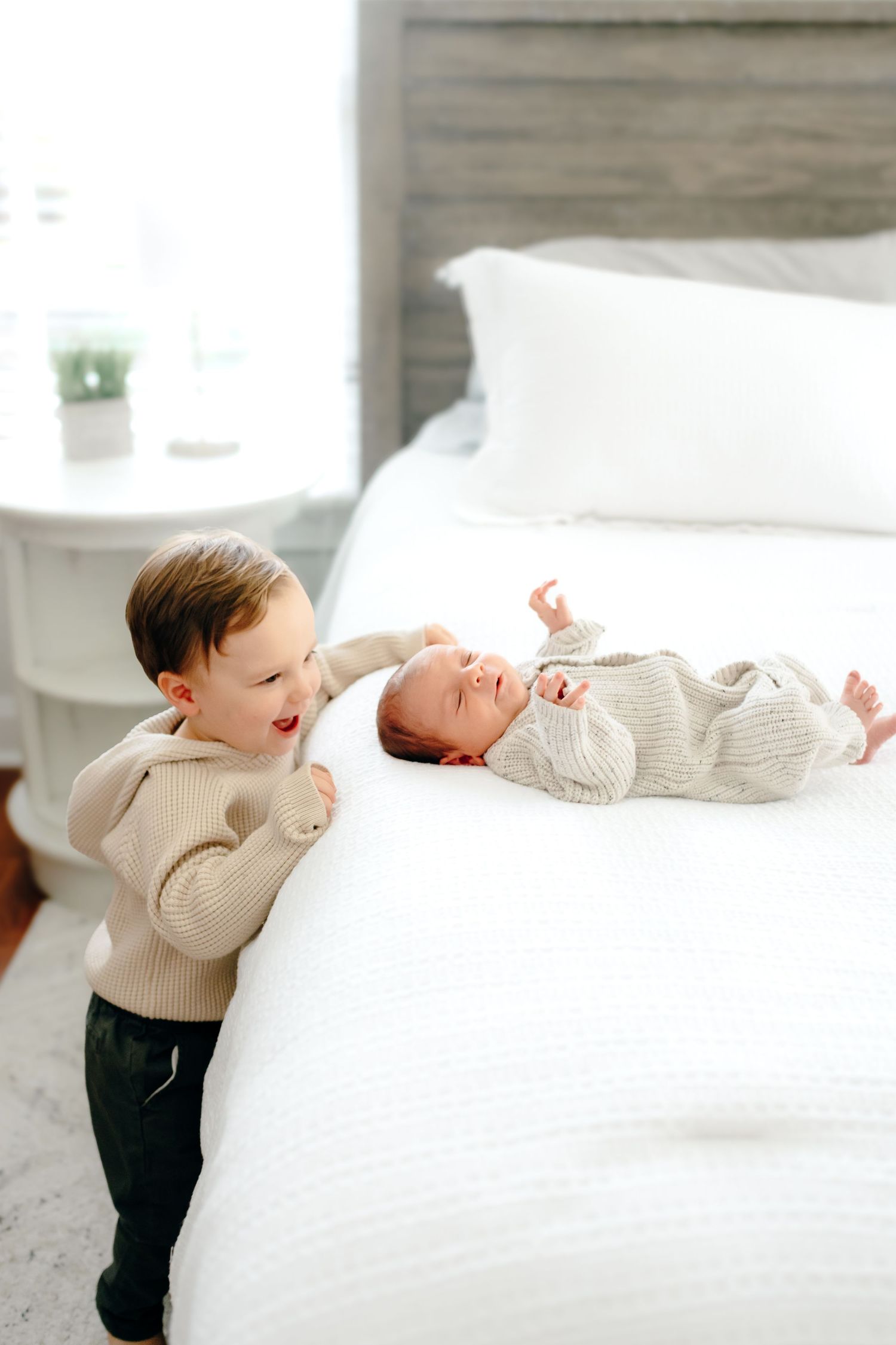 Toddler in beige sweater excitedly looks at baby sibling on white bed.
