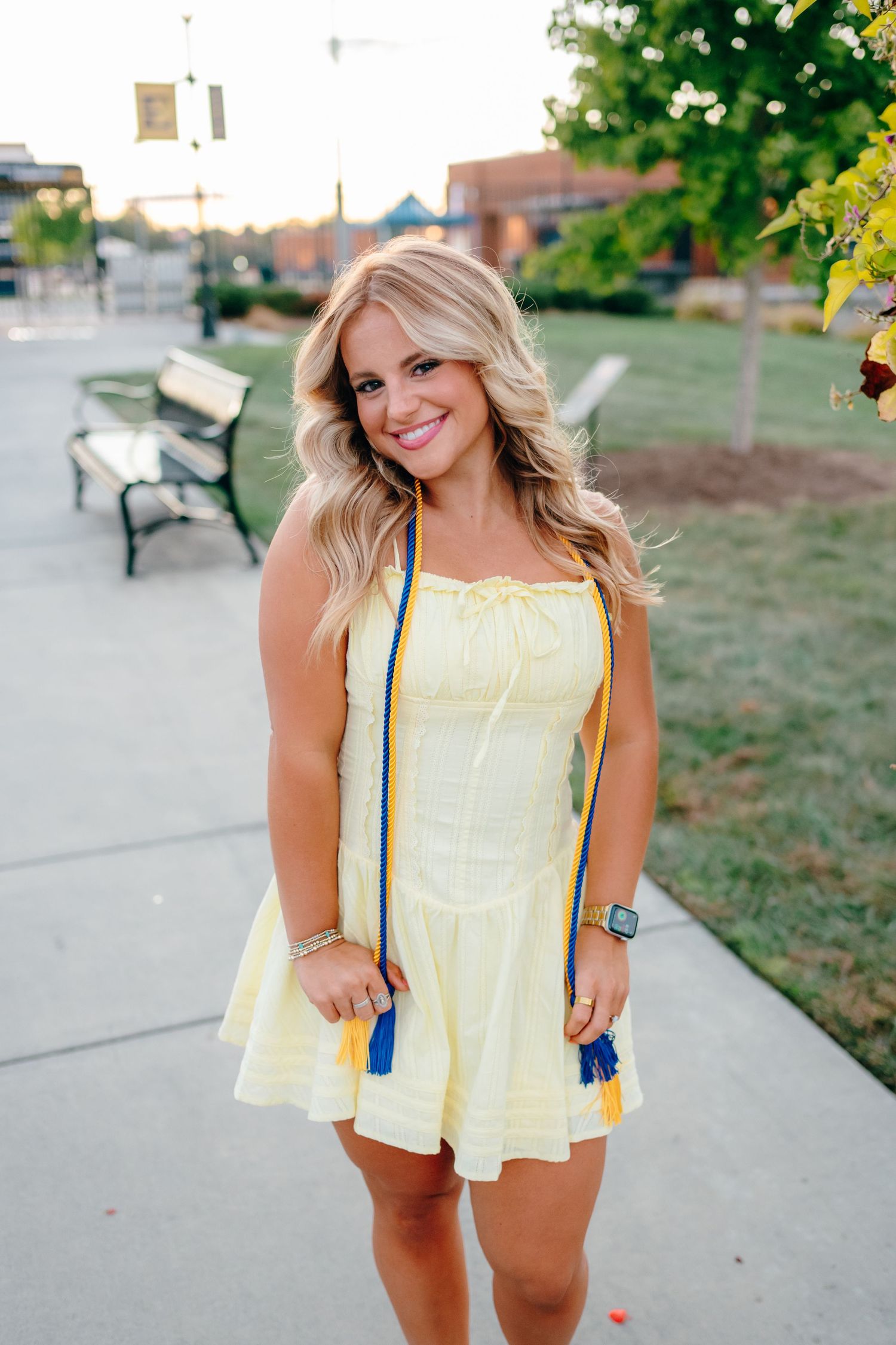 Graduate in yellow sundress poses on campus pathway during sunset.