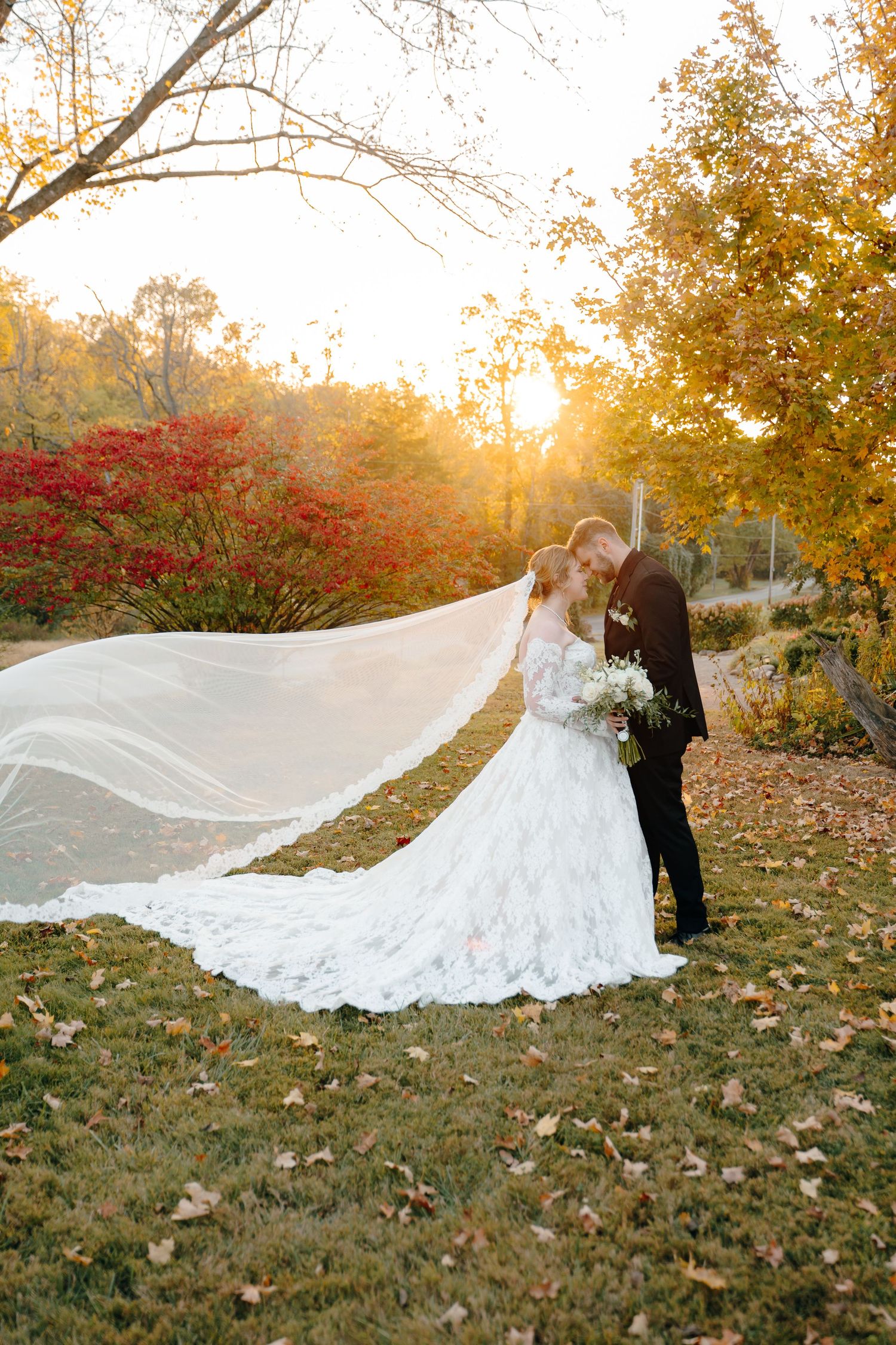 Dramatic wedding photo with flowing cathedral veil against autumn foliage at sunset.