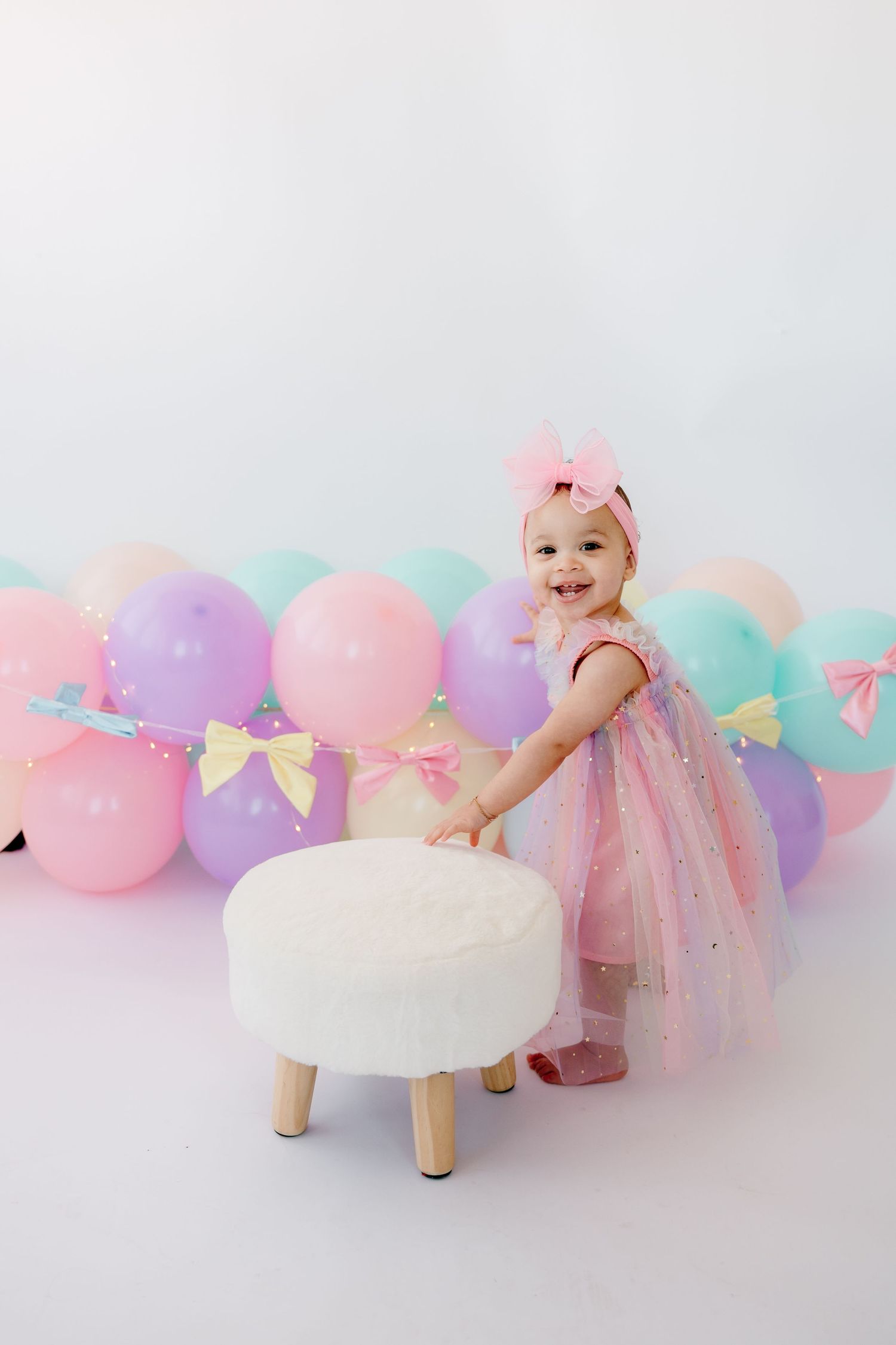 Baby in pink tulle dress playing near pastel balloons and bows against white backdrop.