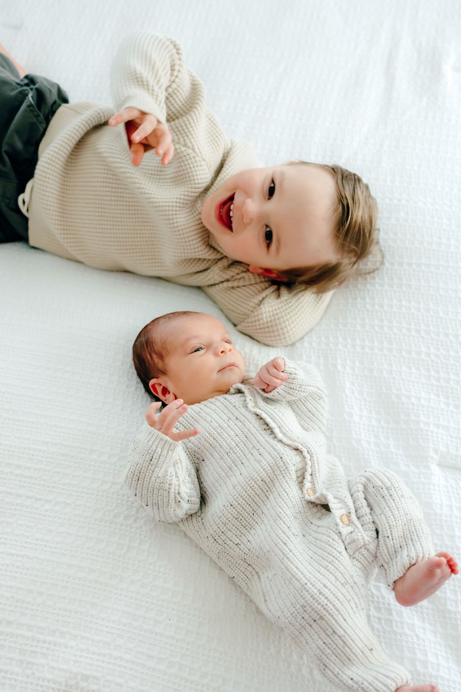 Two children in cozy sweaters lying on white bedding together.
