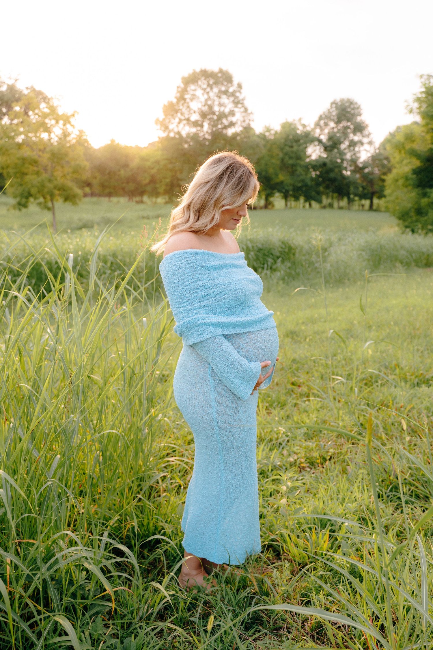 A woman in a light blue dress stands in a sunlit field during golden hour.