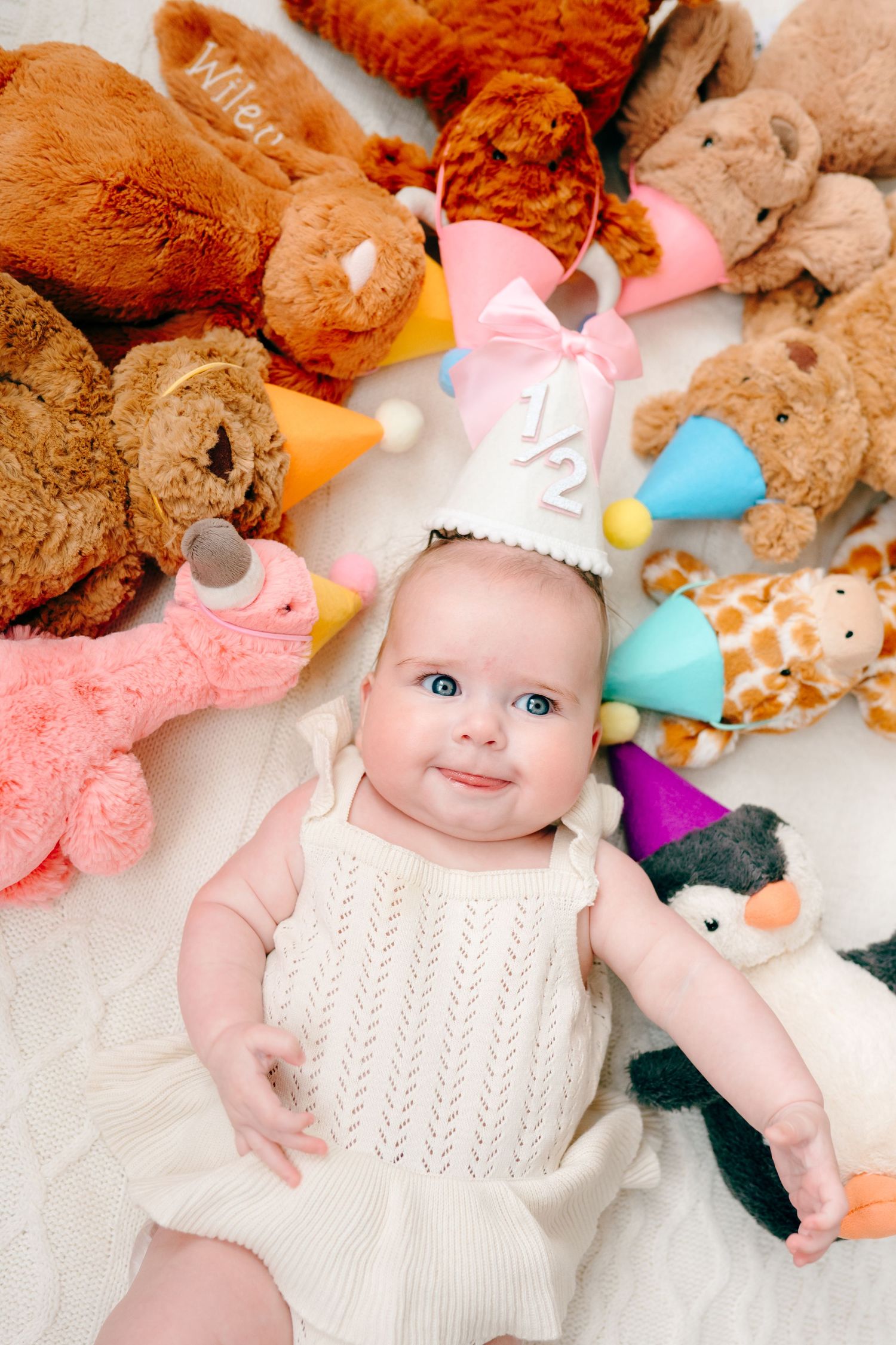Baby in white dress surrounded by colorful plush stuffed animals on white background.