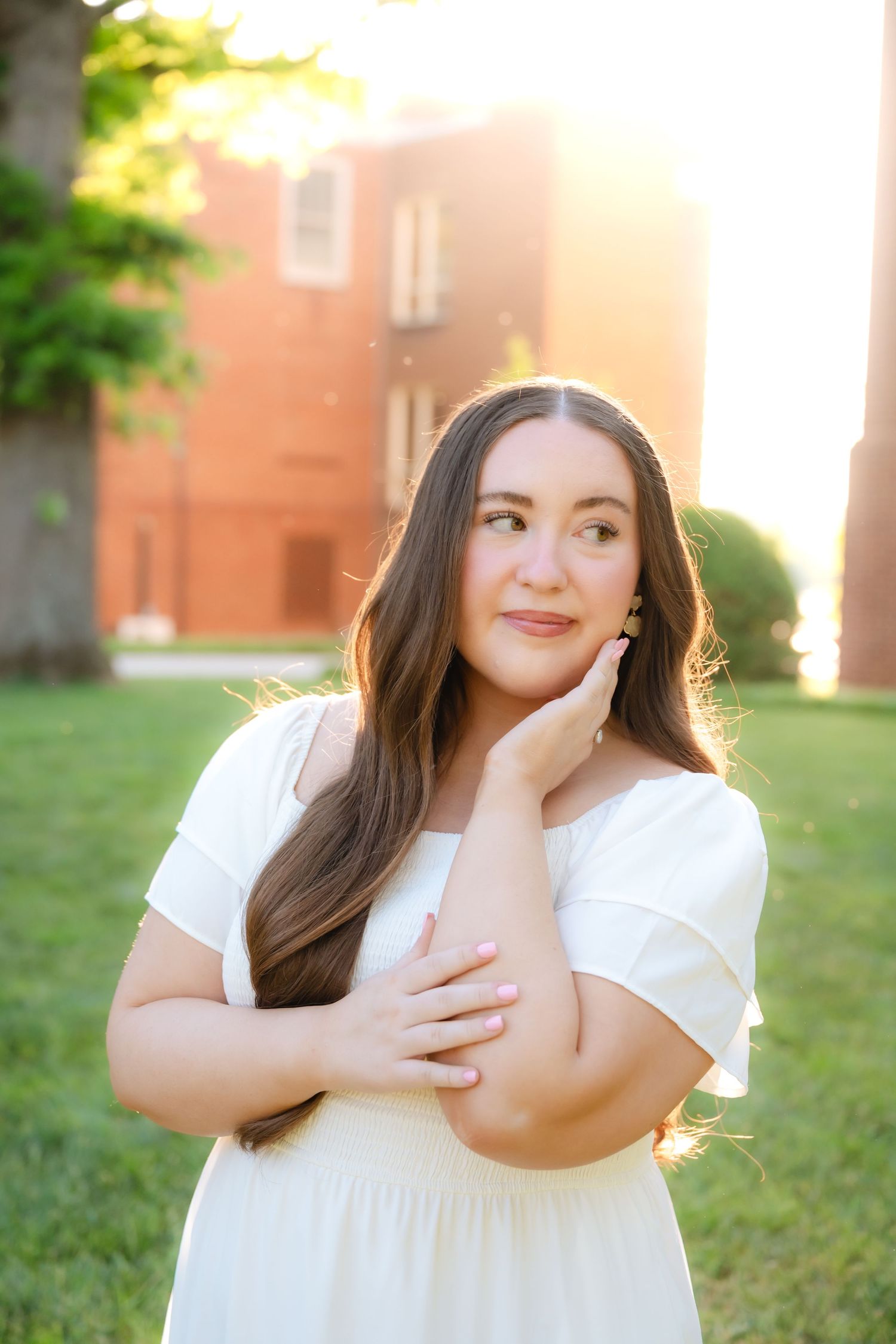 Young woman in white dress poses outdoors at golden hour near brick building, hand on cheek, long brown hair.