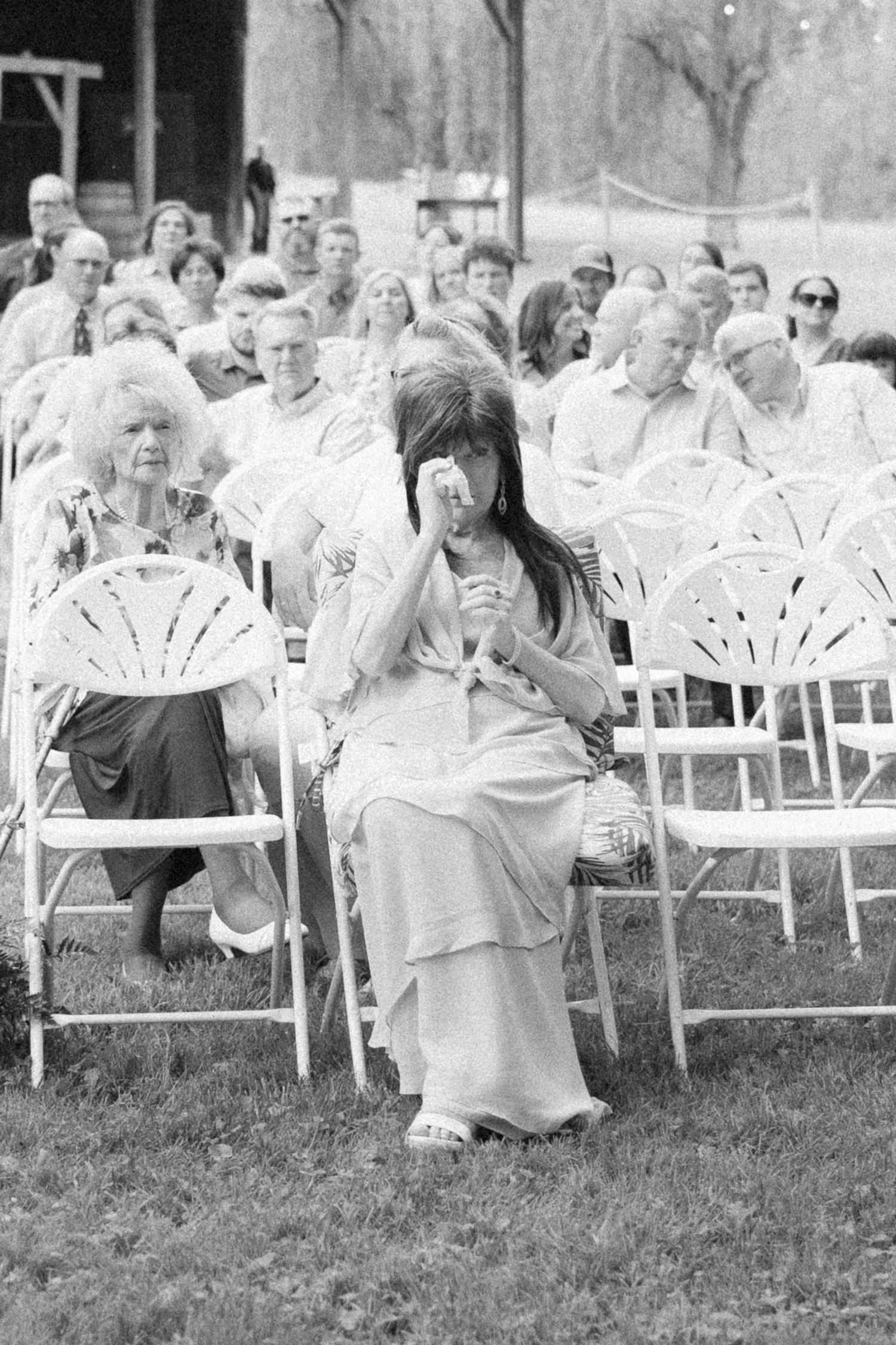 Wedding guests seated in white folding chairs during outdoor ceremony in black and white.