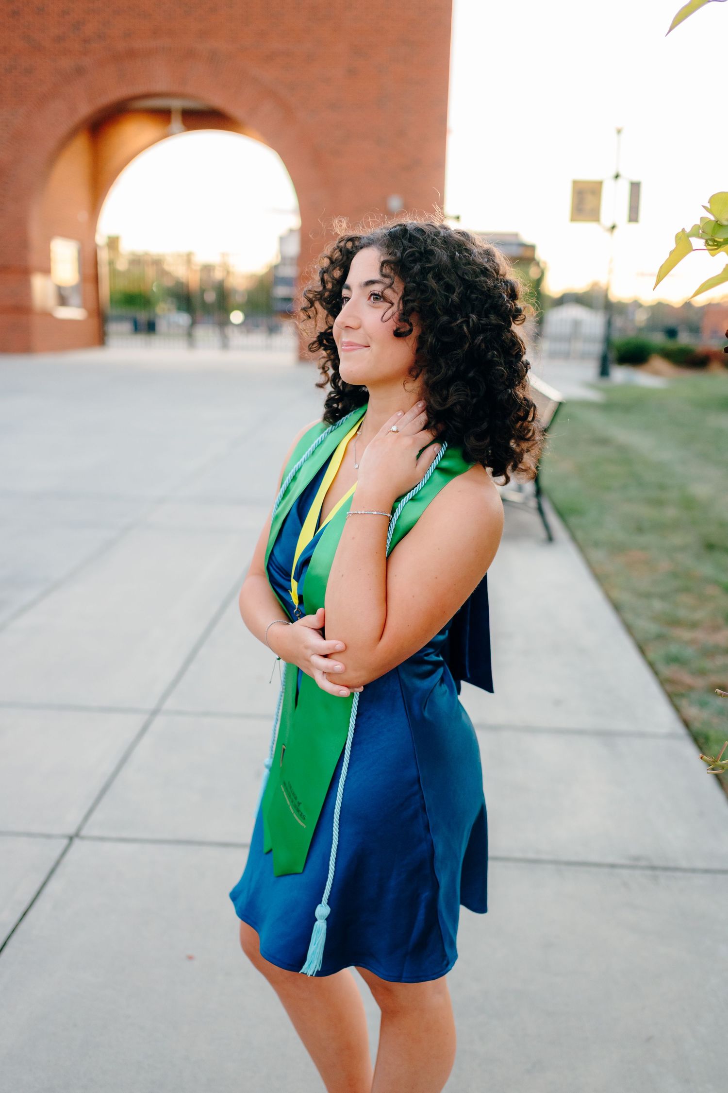 Graduate poses in navy dress and green stole near brick archway during sunset.