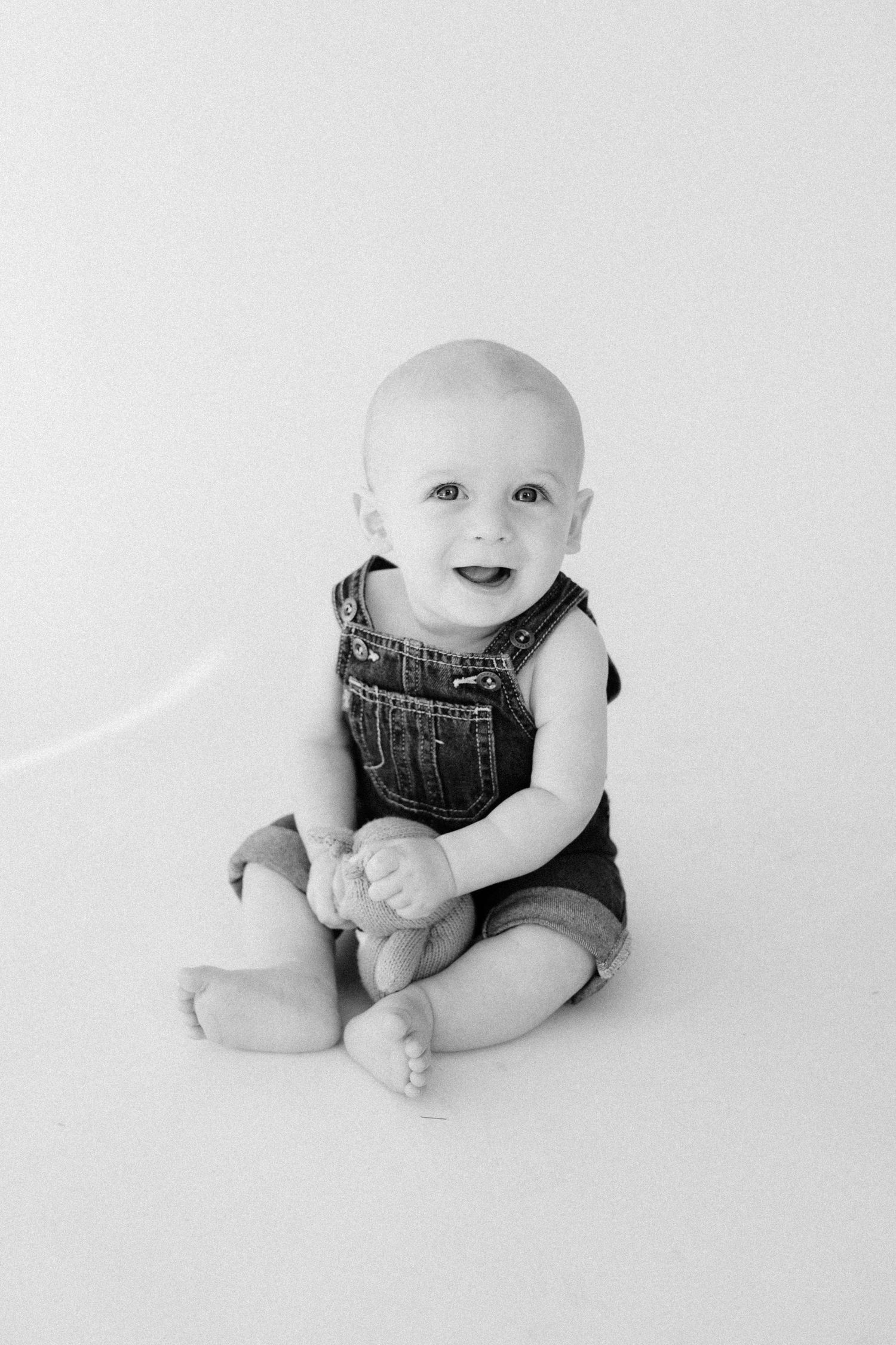 Small baby wearing denim overalls lies on white background in a series of minimalist black and white portraits.