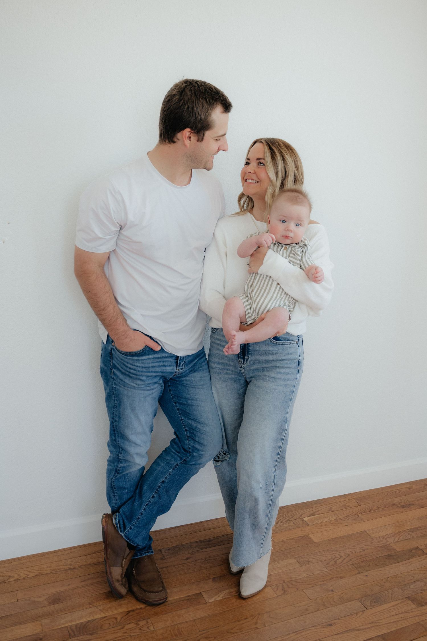 Family photoshoot against white wall showing playful poses in casual denim and white outfits.