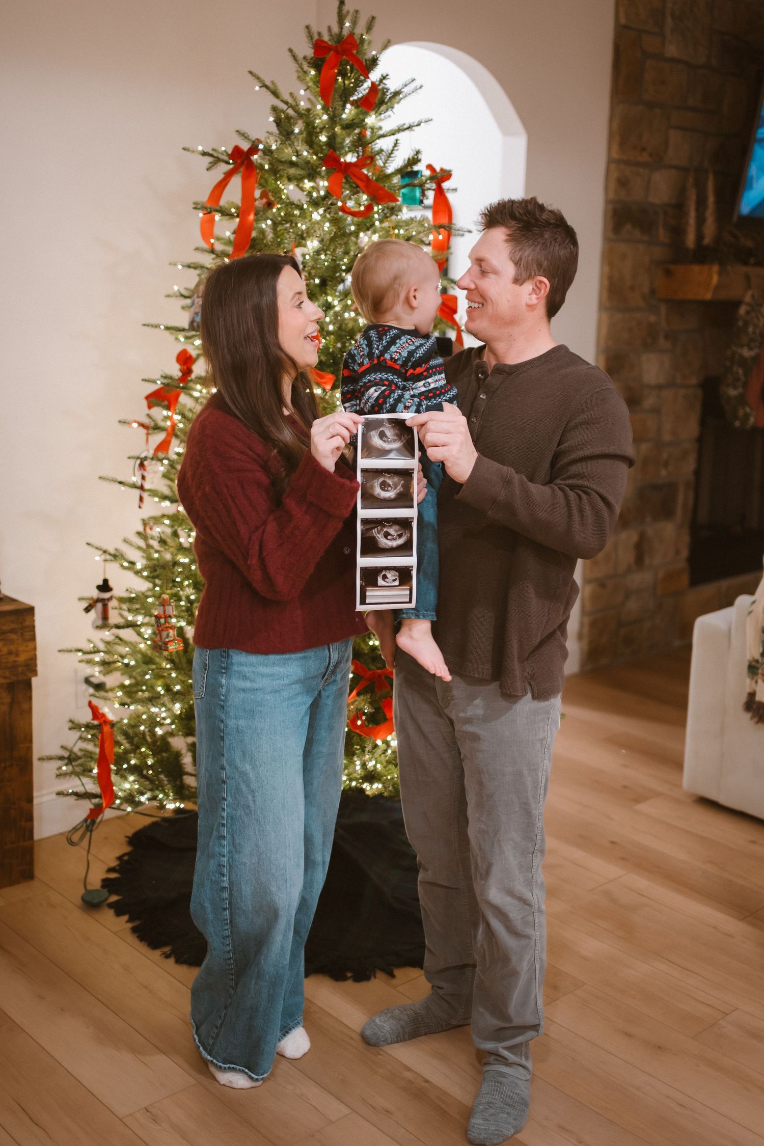 Family celebrates pregnancy announcement with ultrasound photos in front of lit Christmas tree.