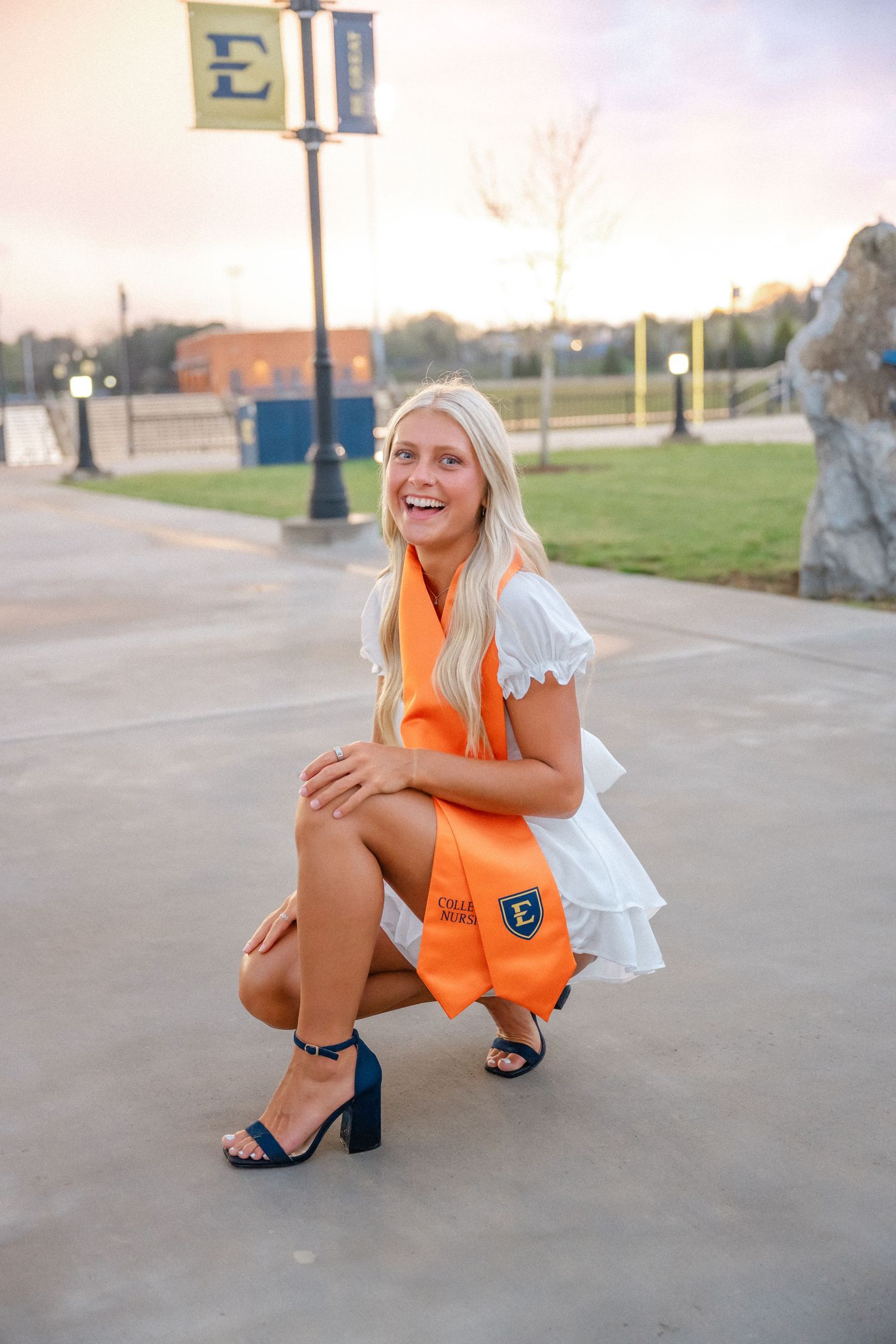 Smiling blonde woman crouching in white dress with orange graduation stole on campus at ETSU.