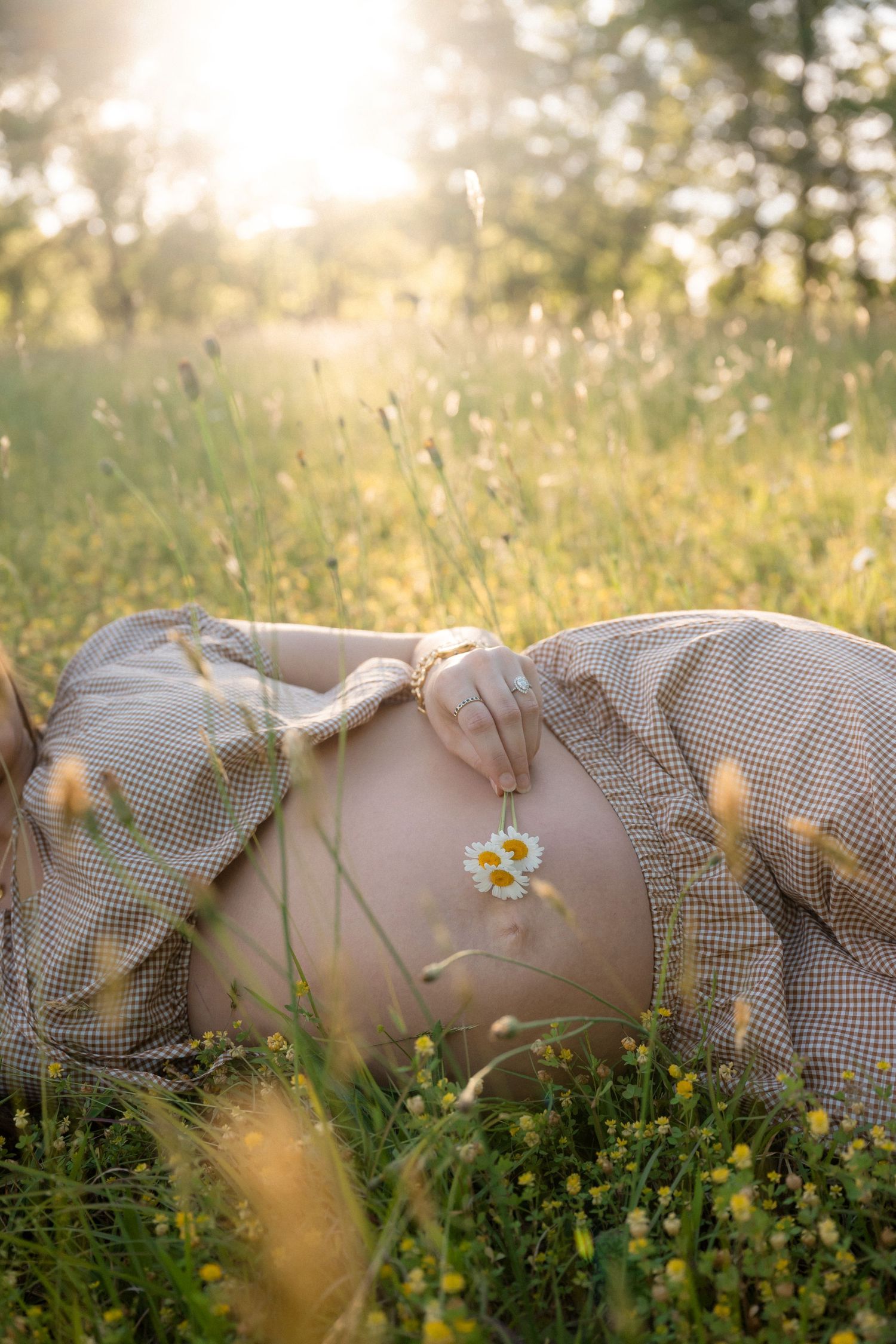 Pregnant woman lying in sunny meadow holding daisy flower on bare belly, golden hour light filtering through green grass.