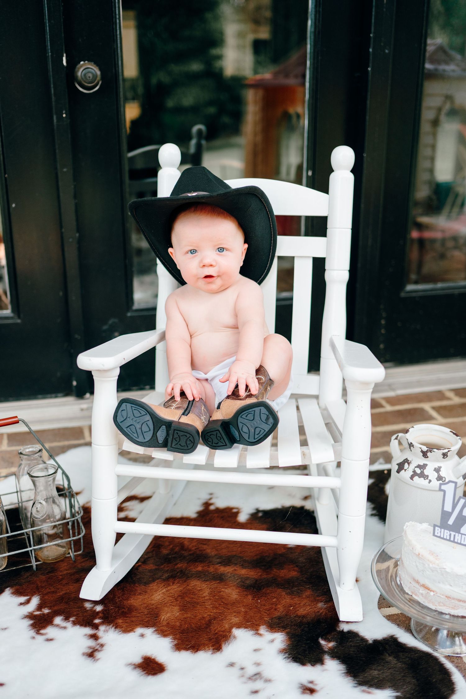 A baby wearing a black cowboy hat sits in a white rocking chair on a porch with a cowhide rug below.