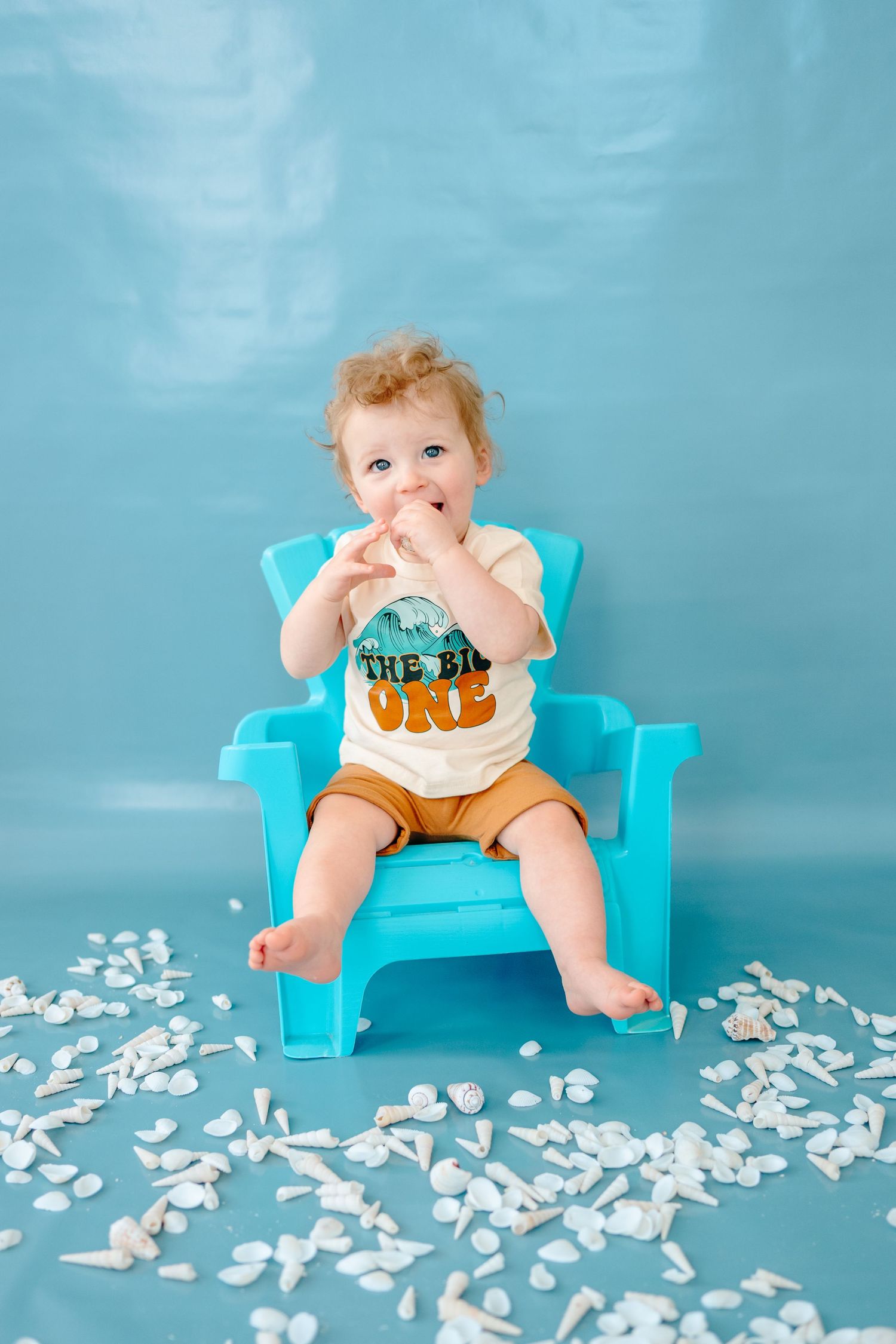 Baby in turquoise chair sits among scattered white paper while wearing white and orange shirt.
