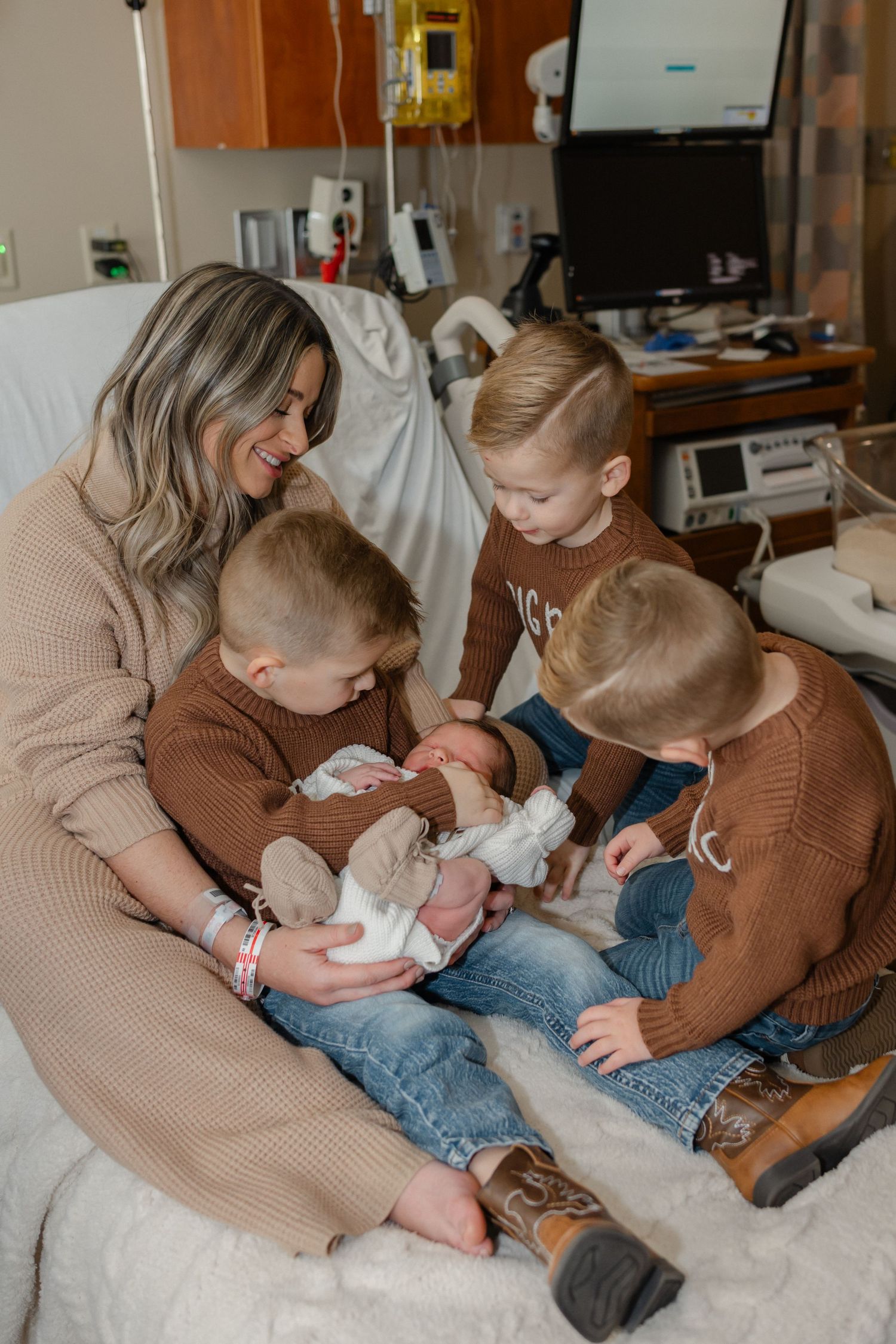 Family sits together on hospital bed while meeting newborn baby in brown sweaters and jeans.