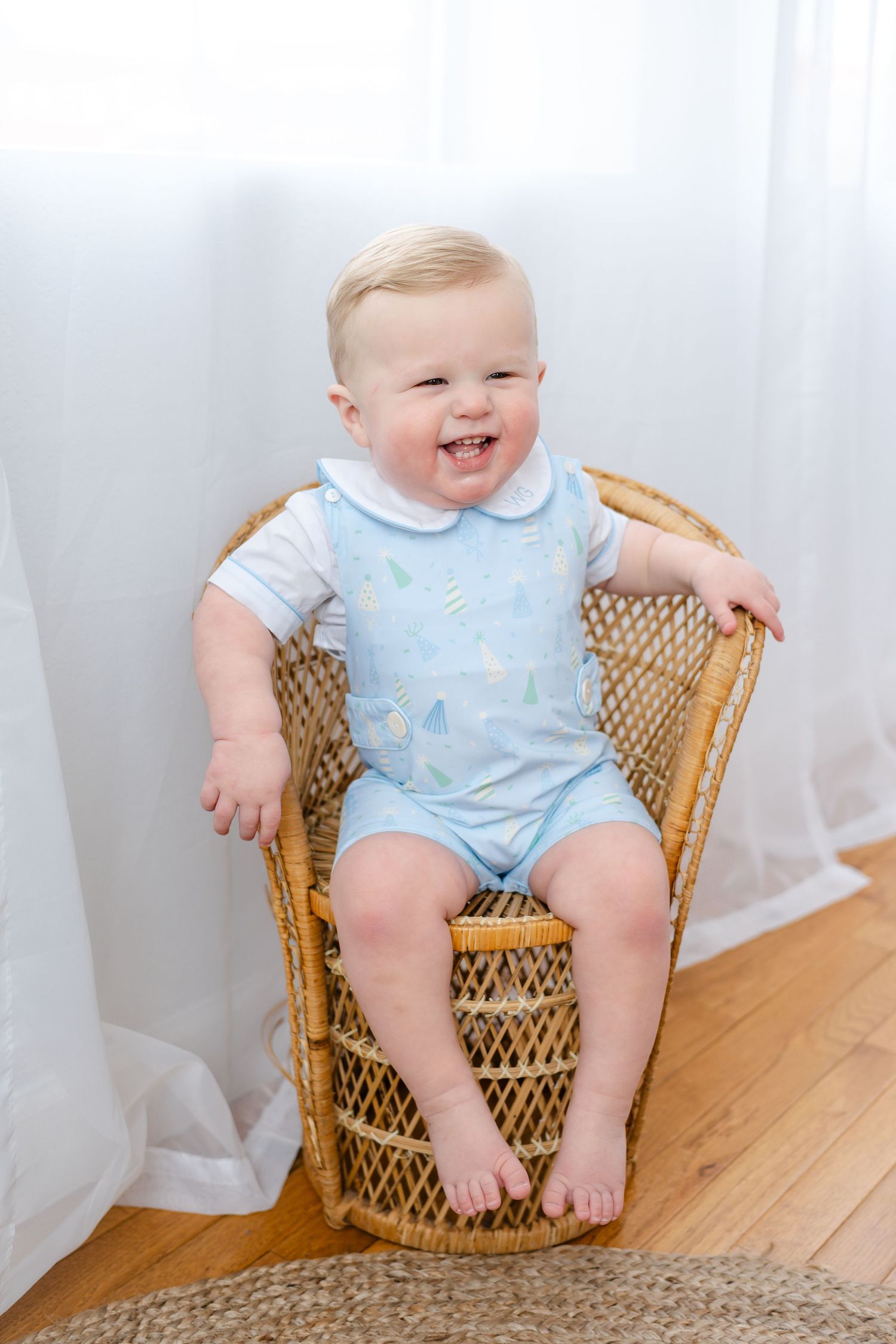 Baby in light blue romper sitting in wicker chair smiles joyfully while posing for milestone photos.