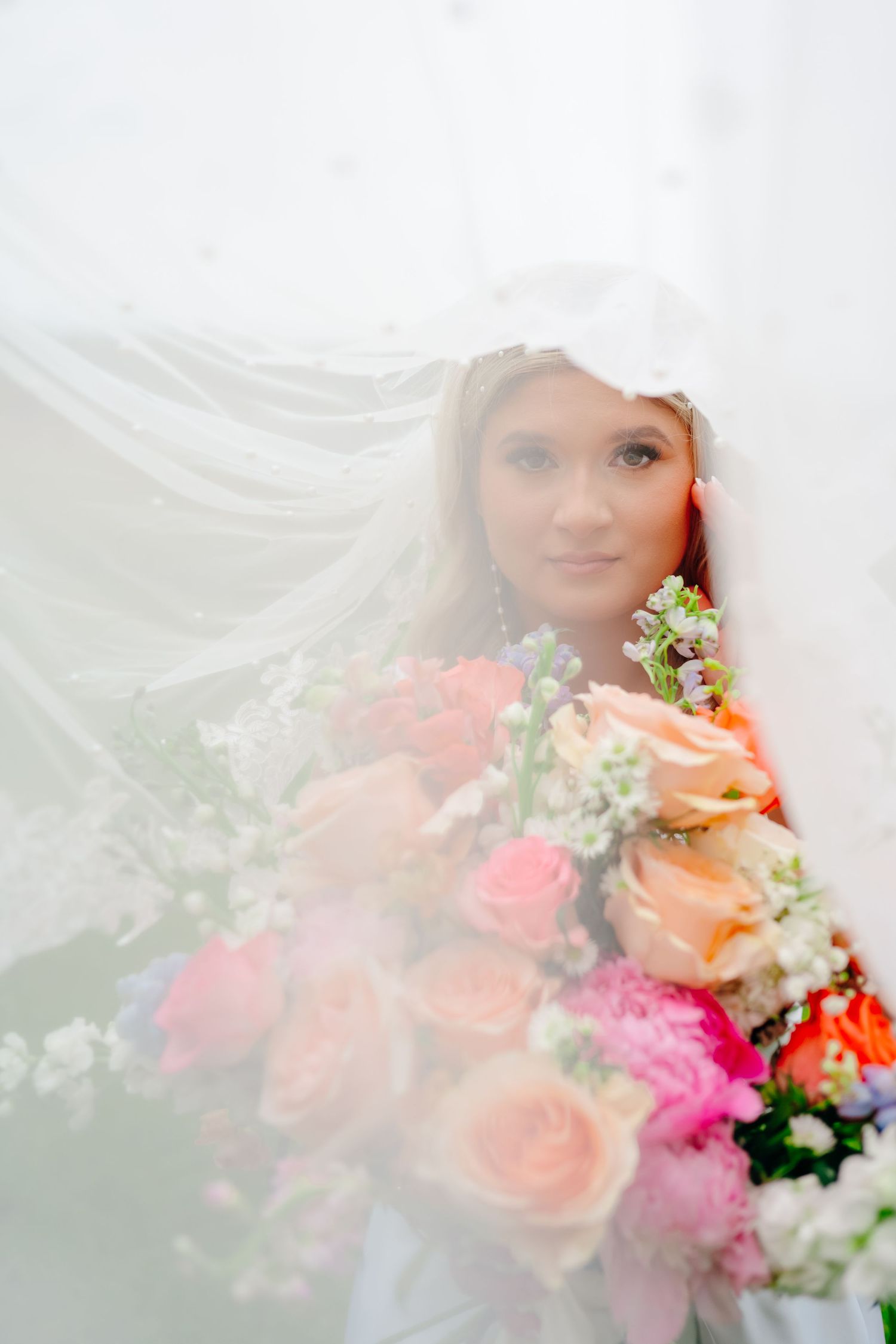 Artistic bridal portrait through veil with coral and pink floral bouquet details.