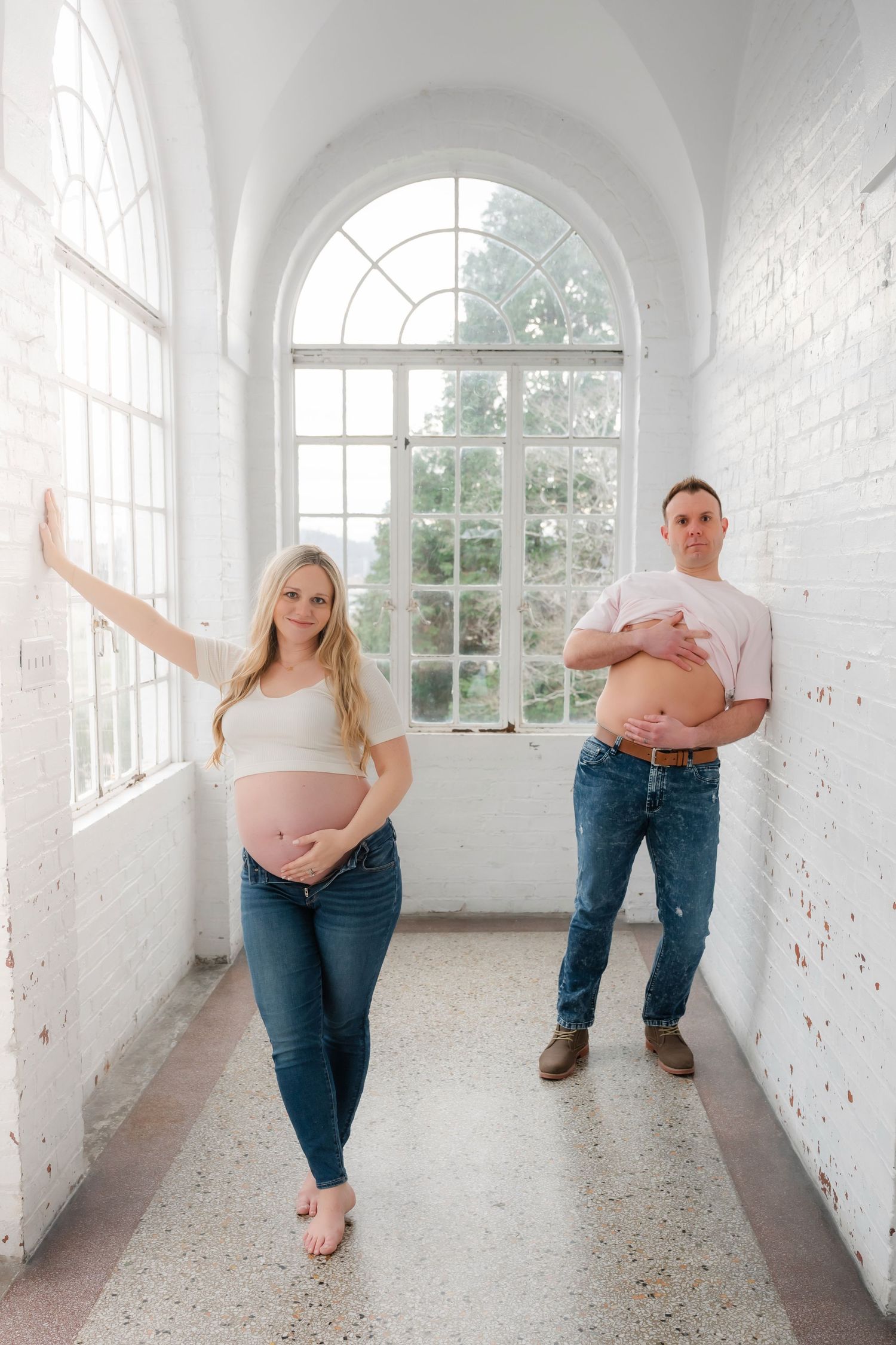 Couple's maternity photoshoot in sunlit white hallway with arched windows.