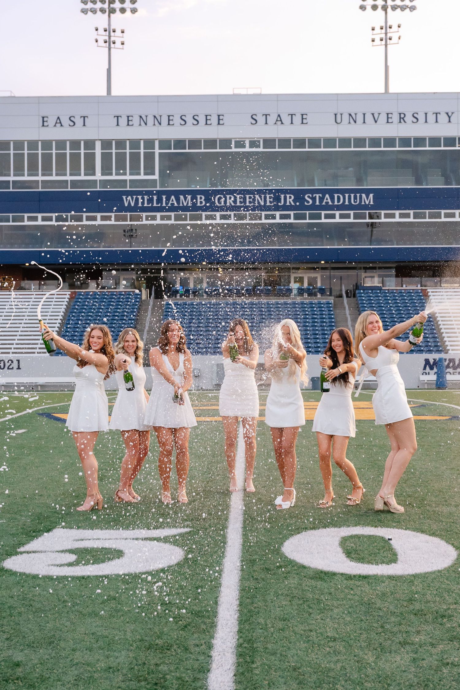 Seven graduates in white dresses spray champagne on East Tennessee State University football field.