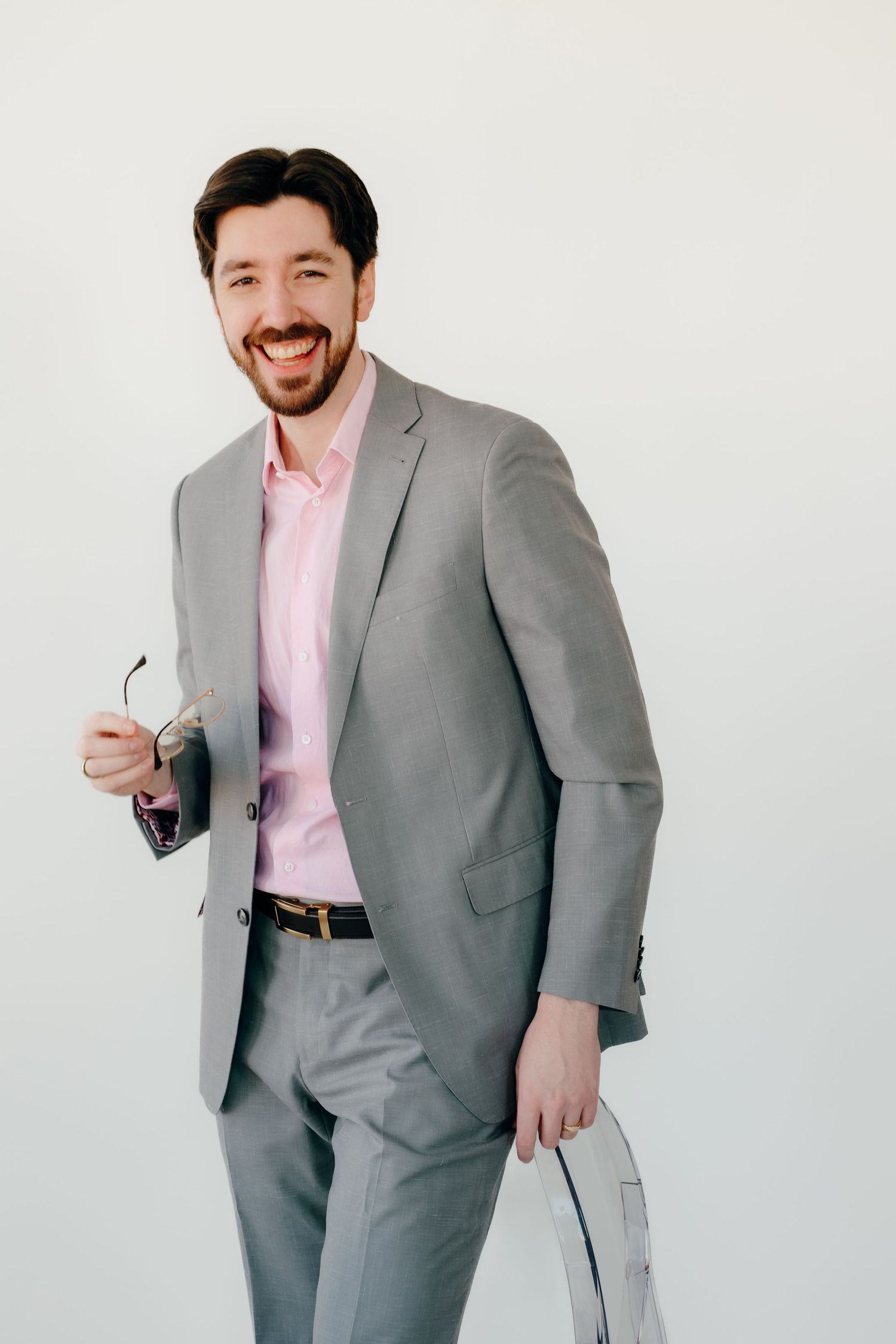 Professional in a light gray suit and pink dress shirt smiling while holding documents against white background.