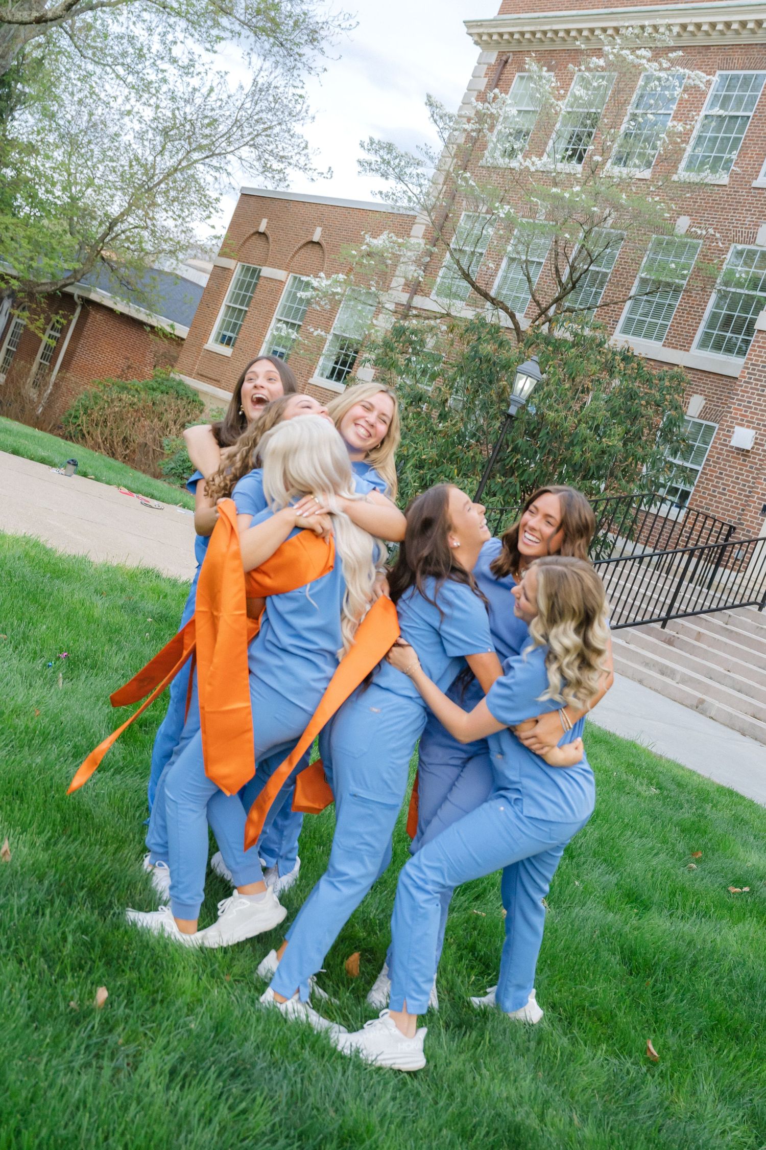 Group of nursing students in blue scrubs celebrating outdoors on campus green, laughing and hugging joyfully.