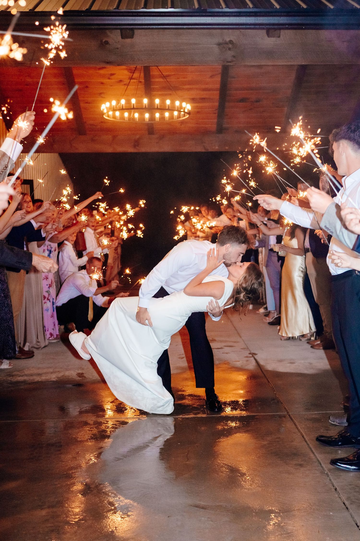 Romantic wedding exit with sparklers as couple performs dramatic dip kiss under rustic chandelier.