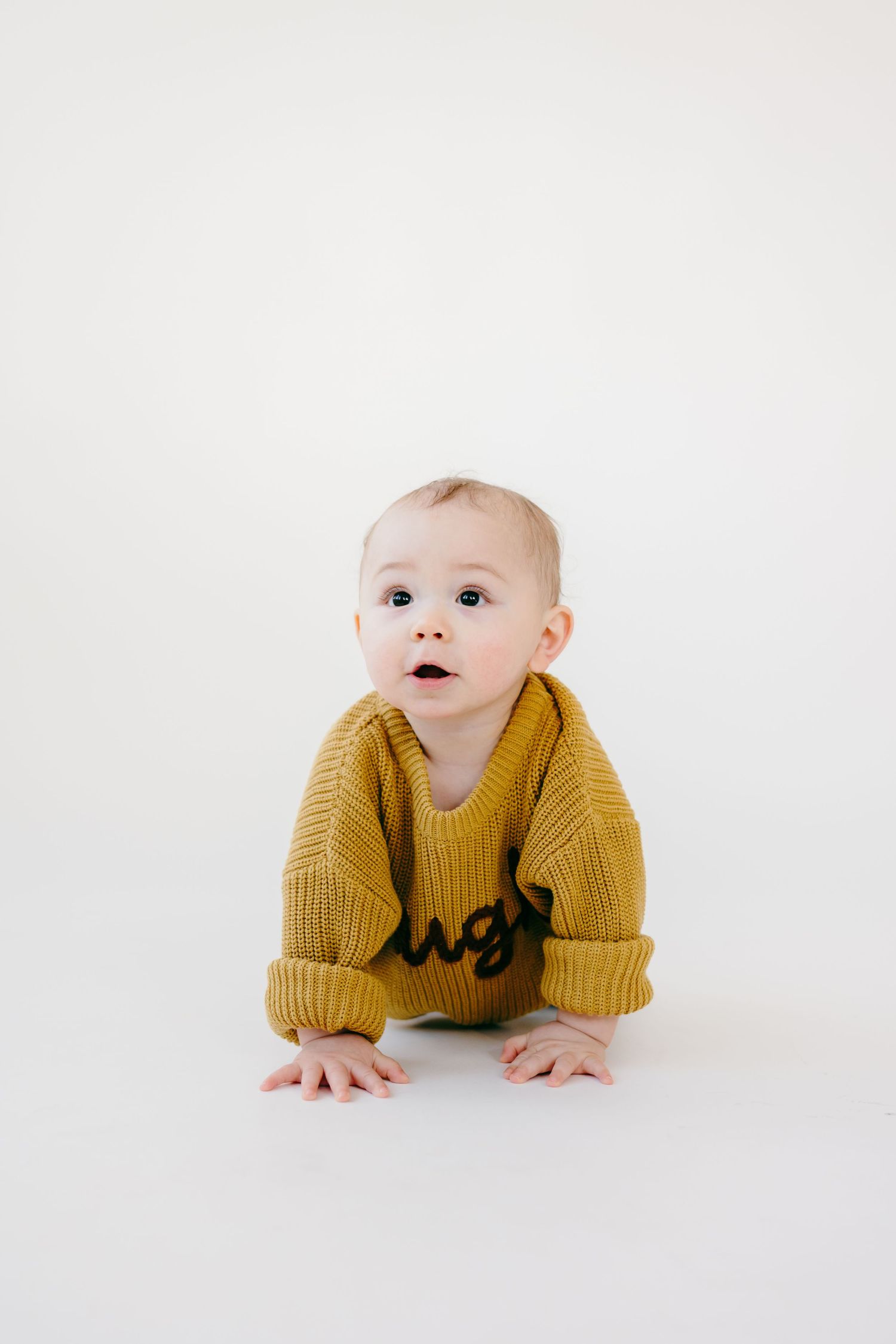 Baby in yellow sweater crawls on white background during photo session.