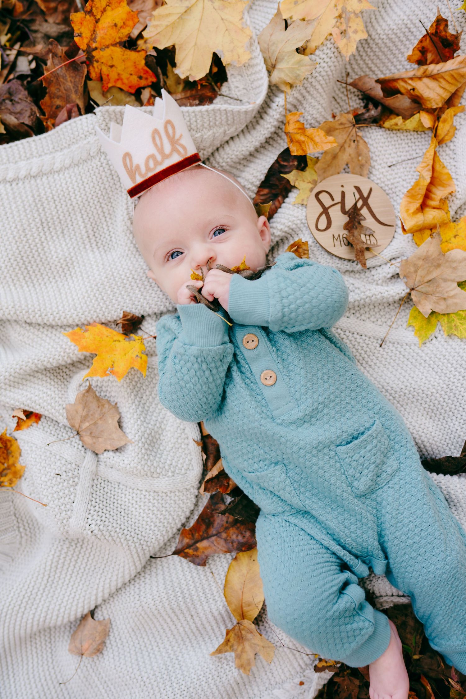 Baby in blue outfit lying on white blanket surrounded by autumn leaves.