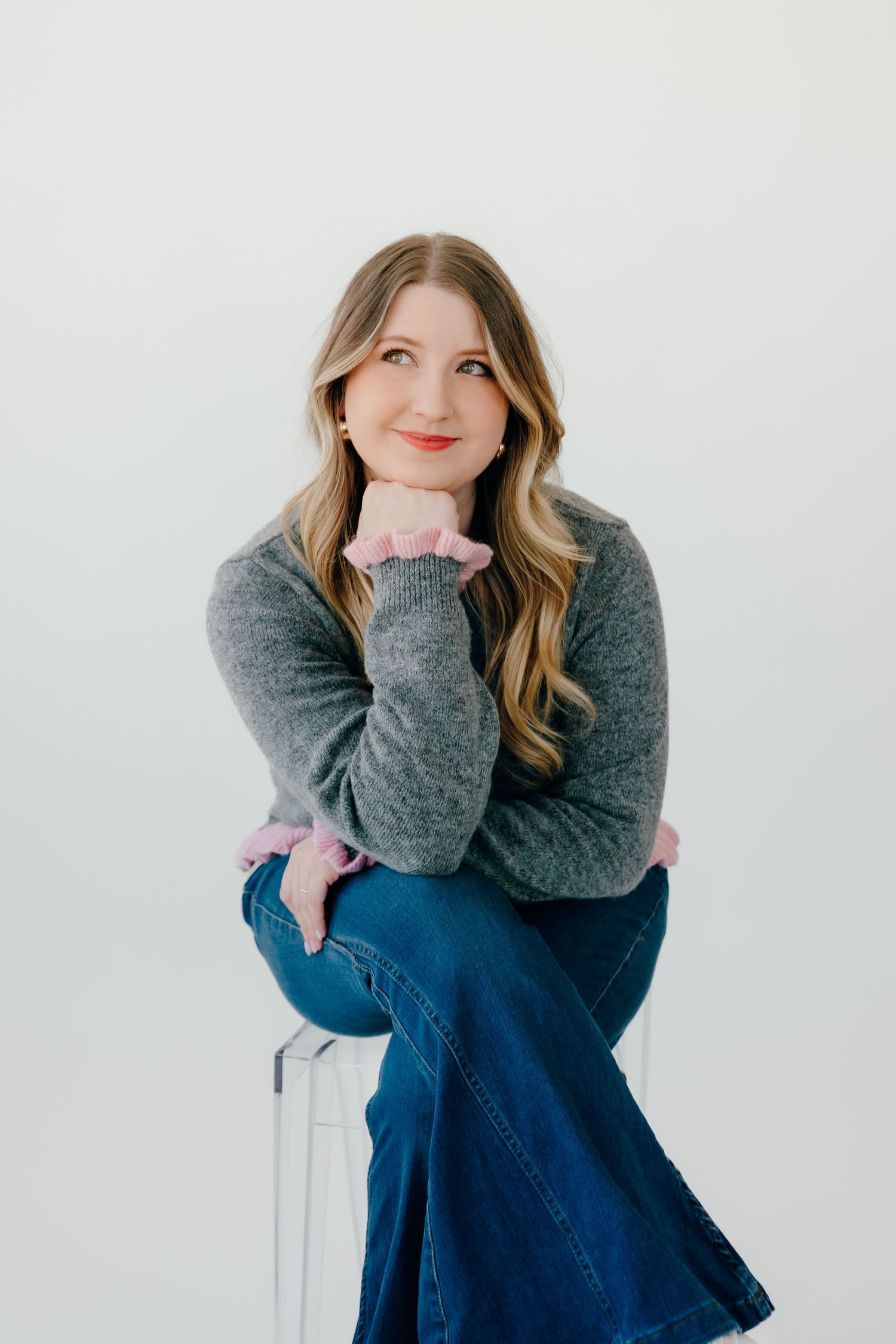 Professional portrait of someone wearing a gray sweater and denim skirt sitting on a white stool against a light background.