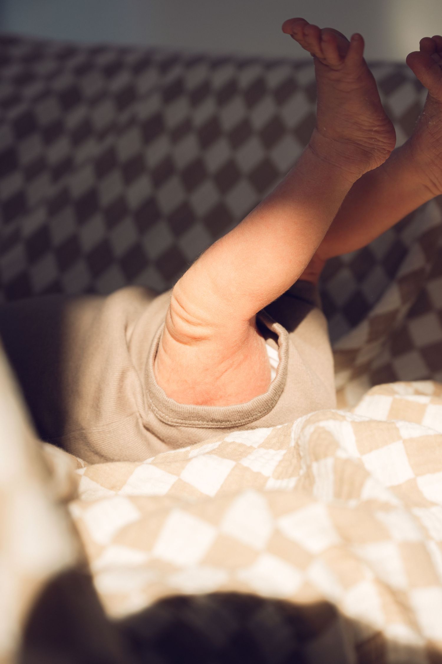 Close-up of a newborn baby's tiny feet and legs in a beige onesie, lying on a checkered blanket.