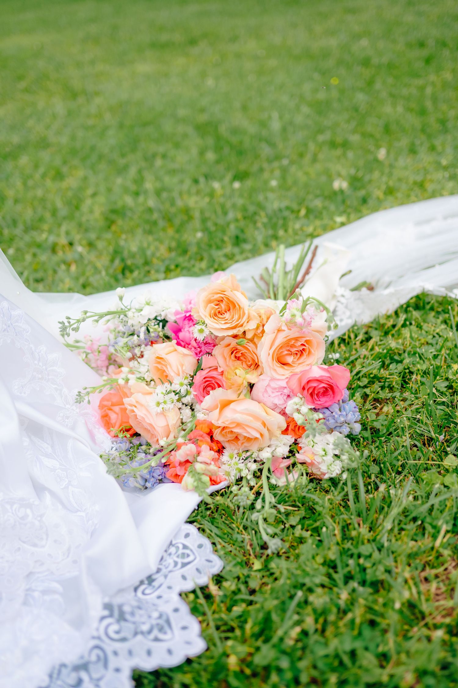 Coral and pink roses arranged with lace veil on green grass.