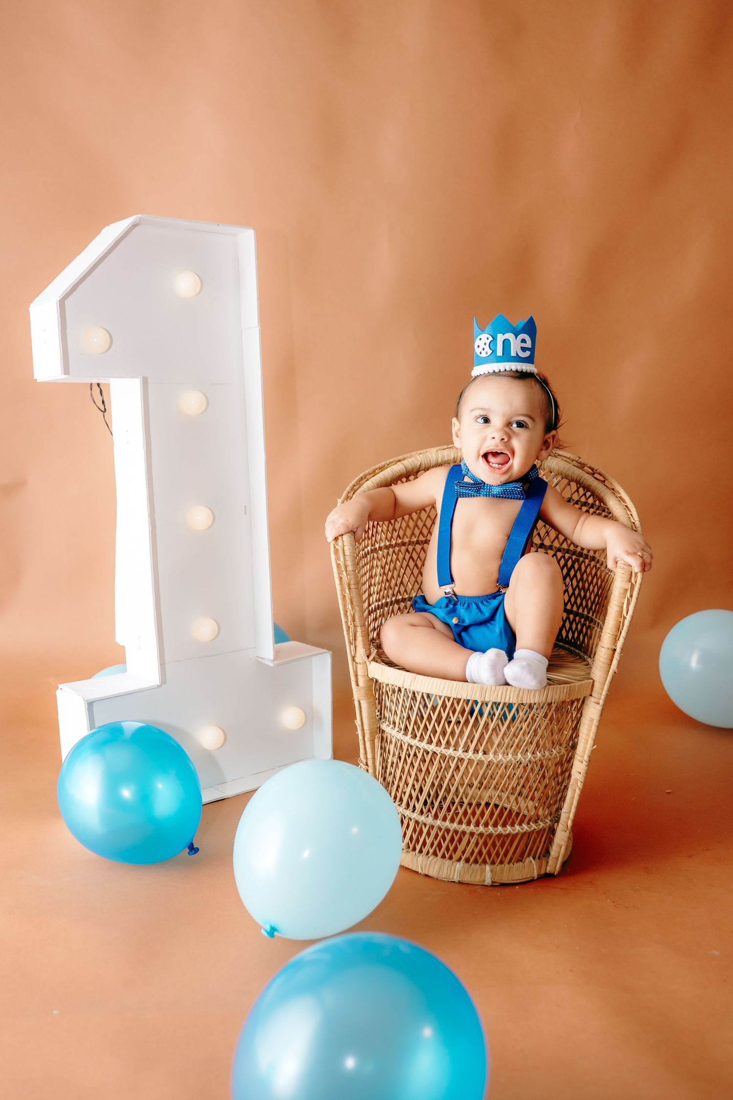 Baby in blue outfit sitting in wicker chair with number one decoration and blue balloons.