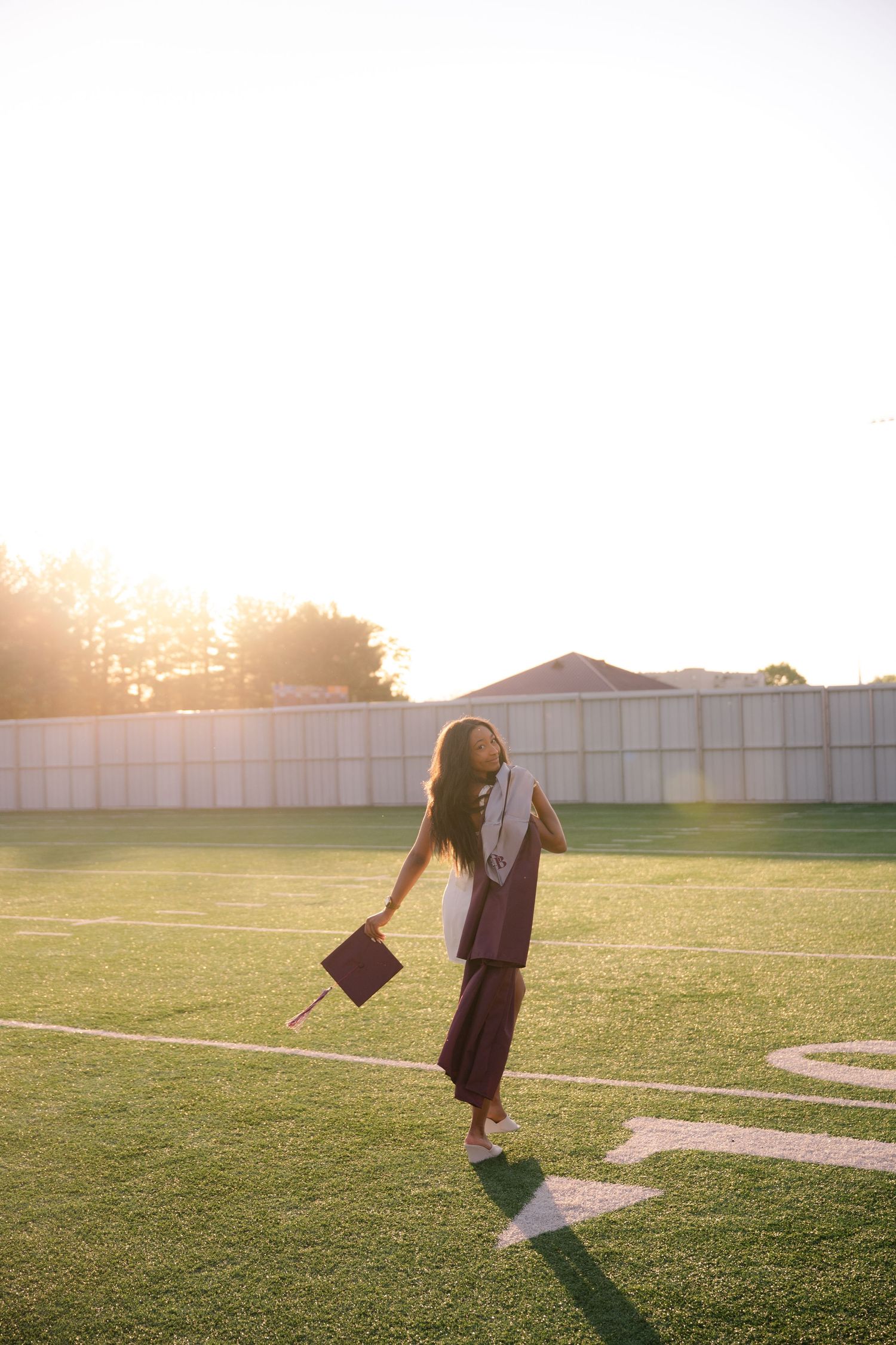Graduate in cap and gown walking on football field at golden hour sunset, holding mortarboard.