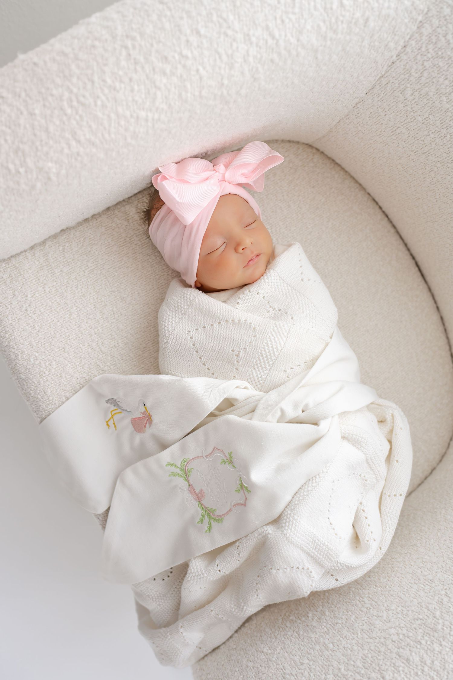 A sleeping baby wearing a white outfit and pink bow rests peacefully on white bedding.