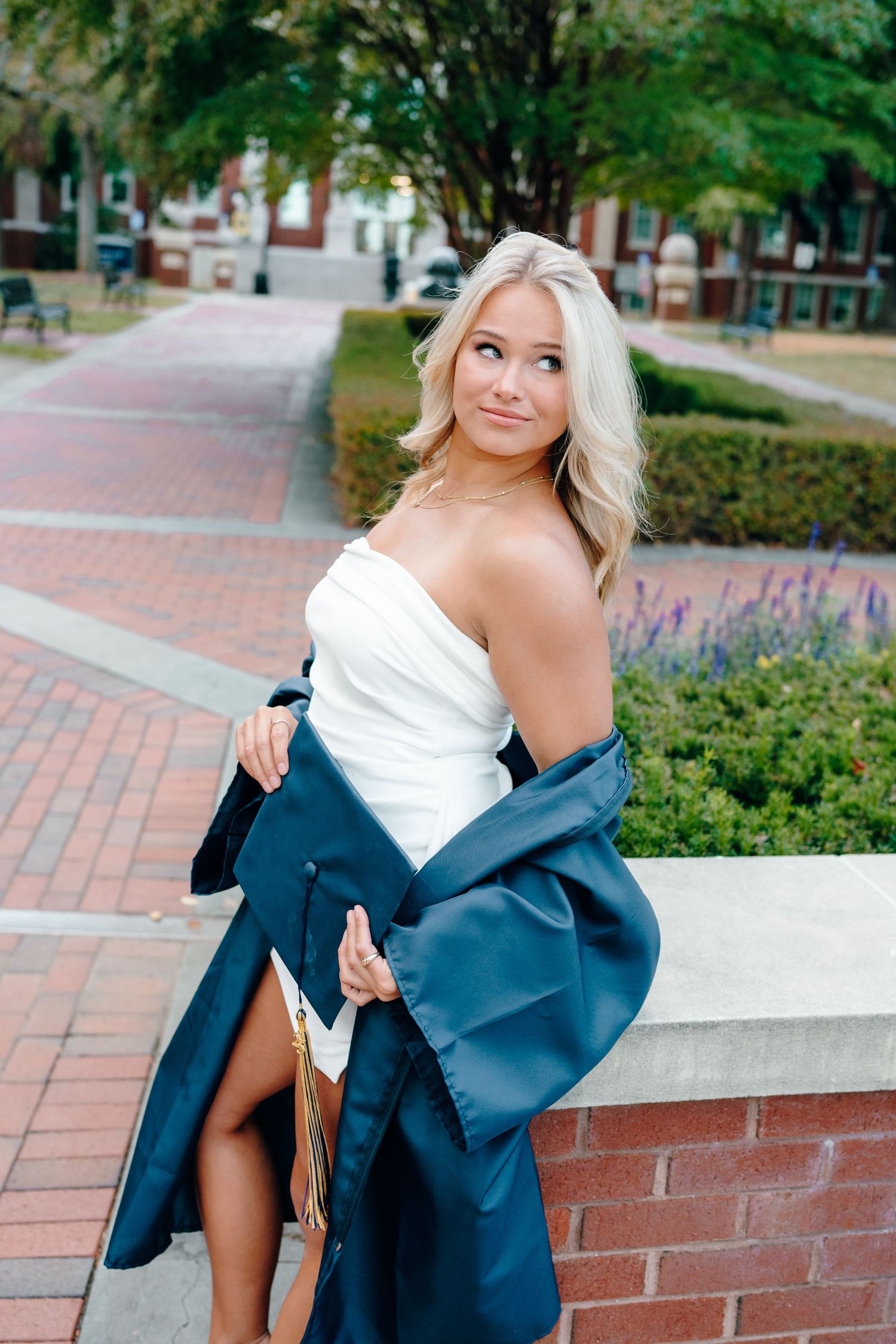 A woman in a white dress and navy blazer poses outdoors on a brick wall near flowering plants and trees.