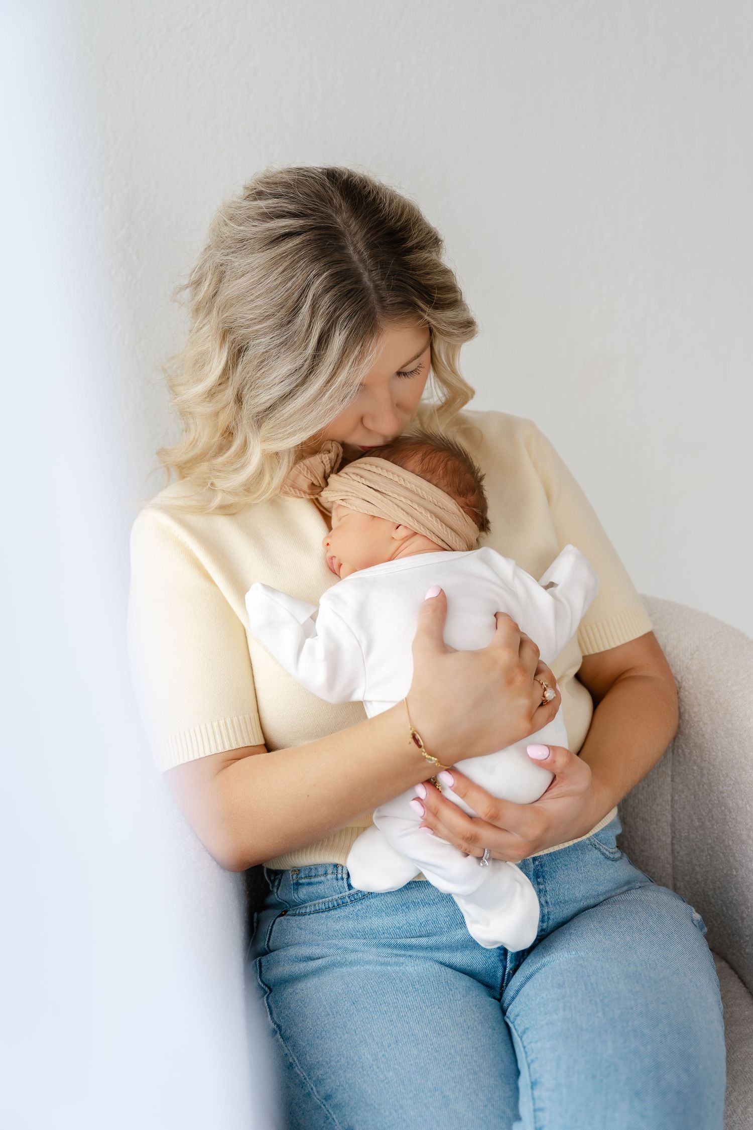 A tender moment between parent and newborn baby dressed in white, sitting against a light gray wall.