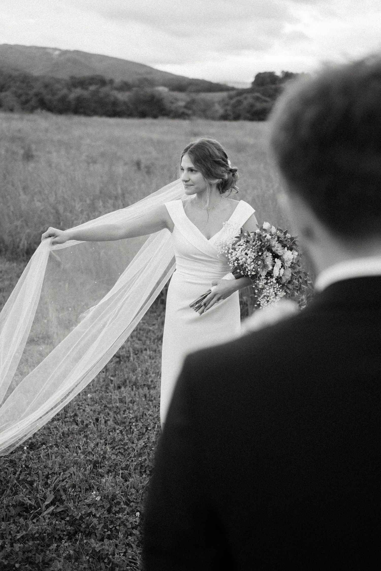 Black and white photo of a bride holding her veil in a meadow.