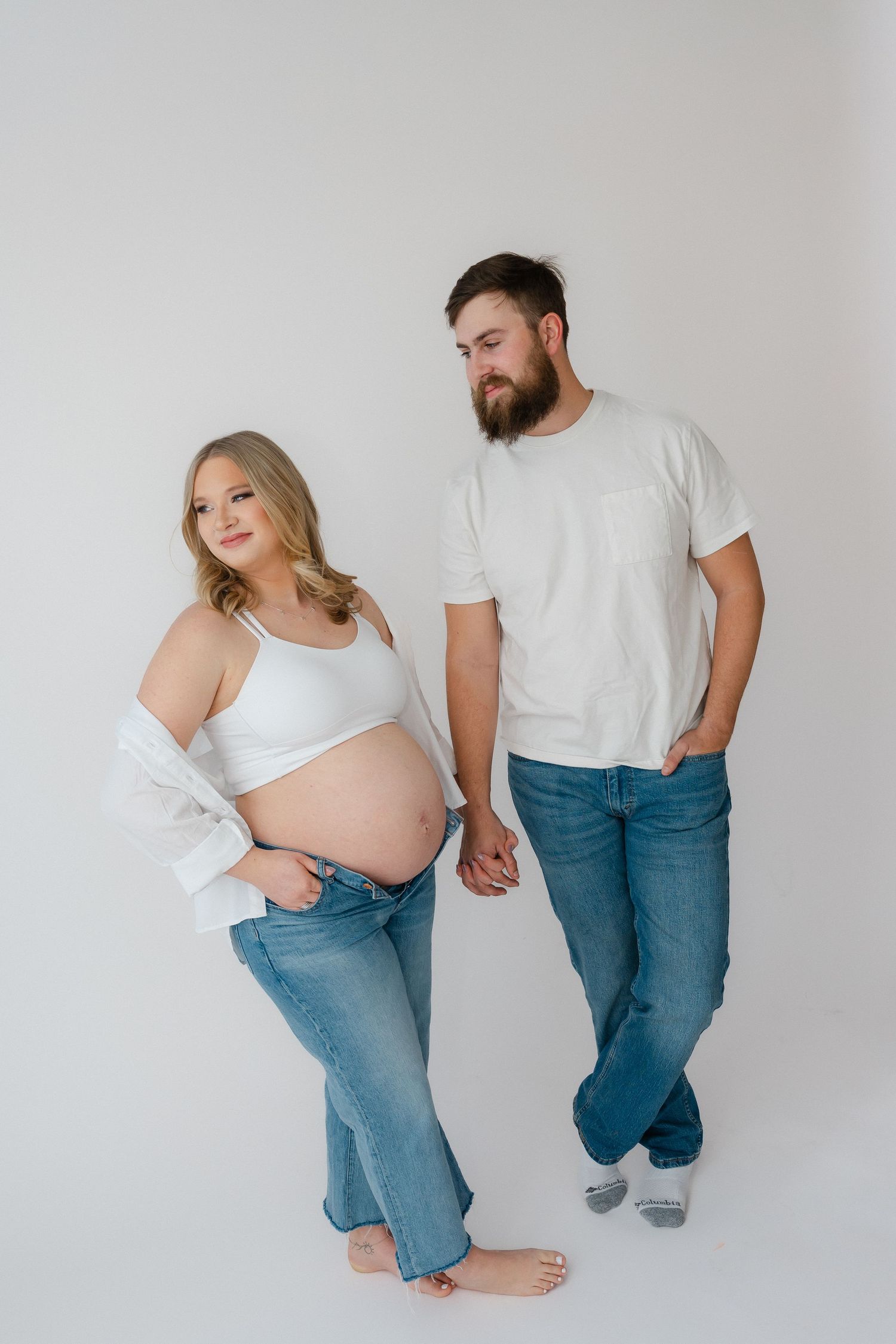 A couple holds hands against a white backdrop while wearing casual denim and white tops in a maternity photoshoot.
