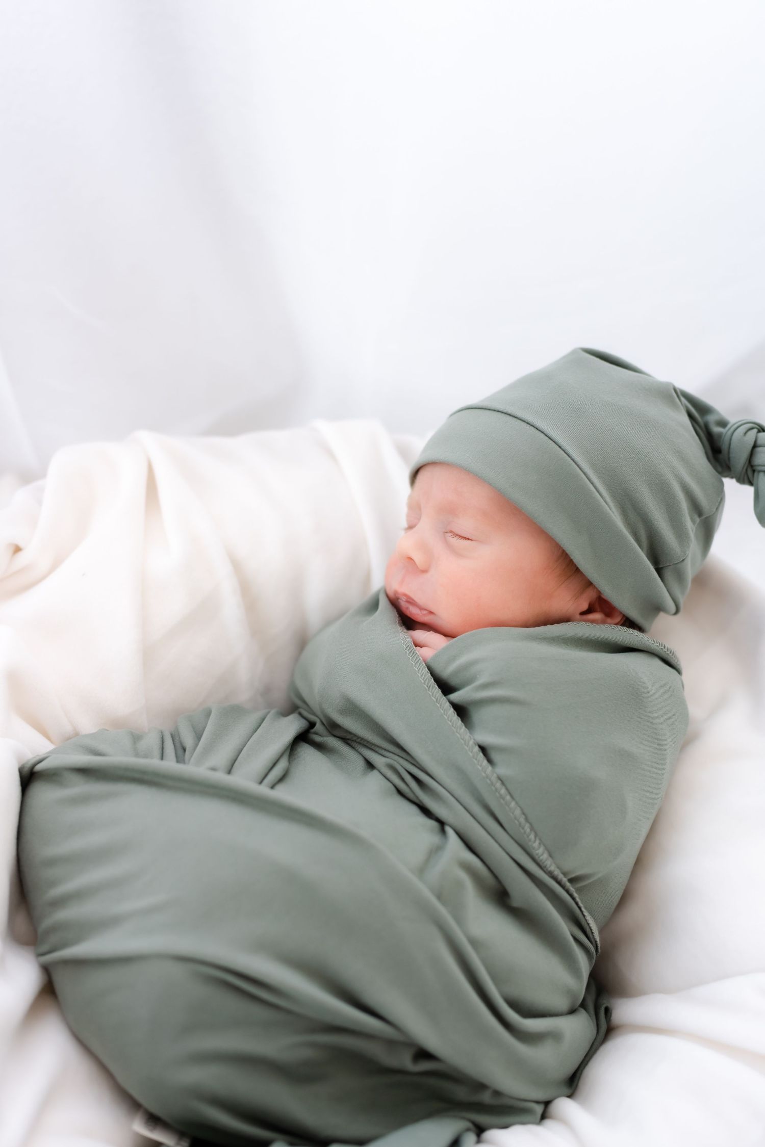 A sleeping newborn baby swaddled in sage green fabric rests peacefully on white bedding.
