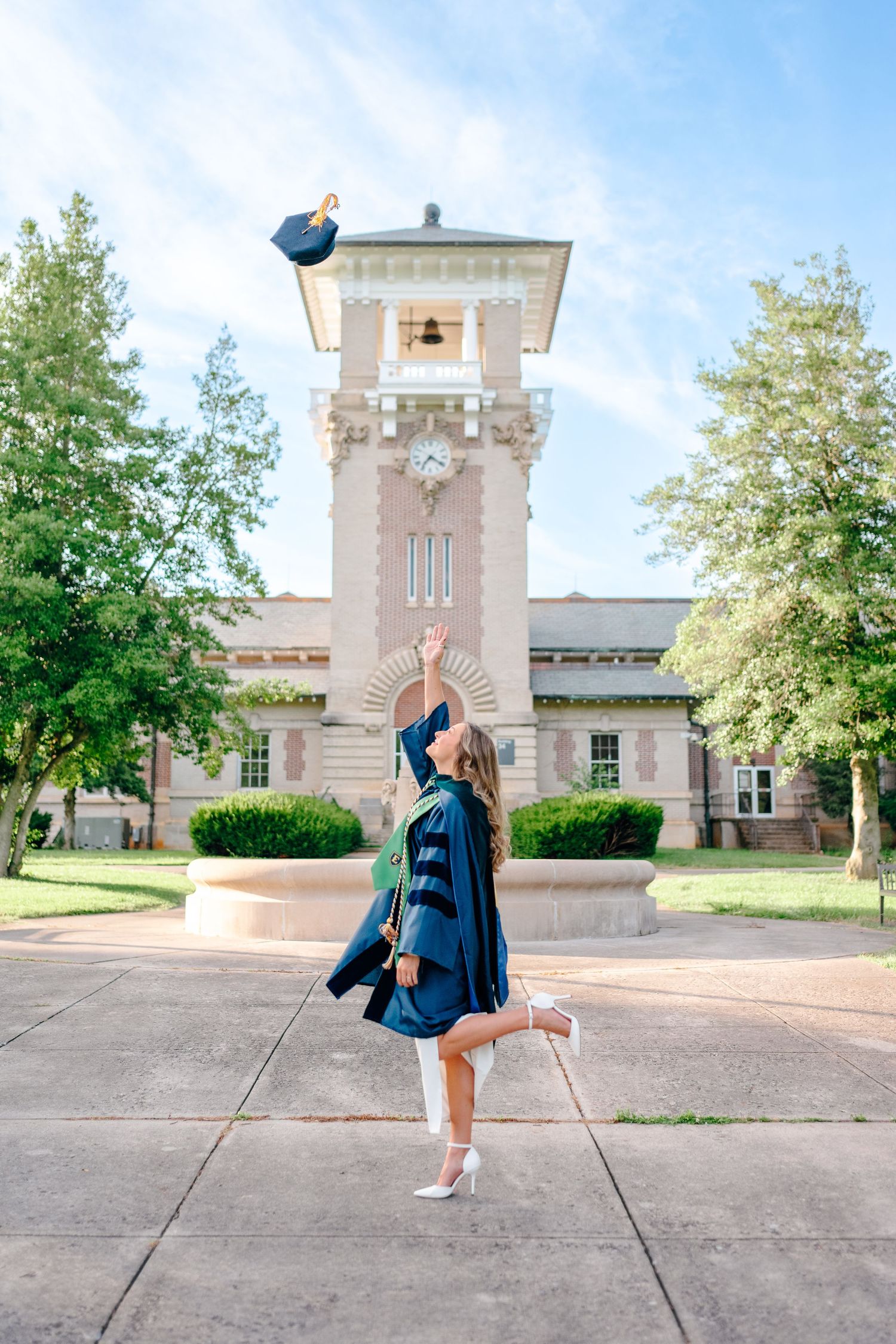 Graduate in academic regalia poses playfully in front of historic campus building.