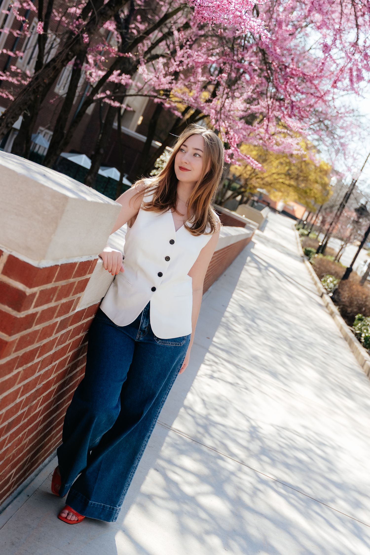 Graduate in white top and jeans poses under blooming cherry blossoms on campus walkway.