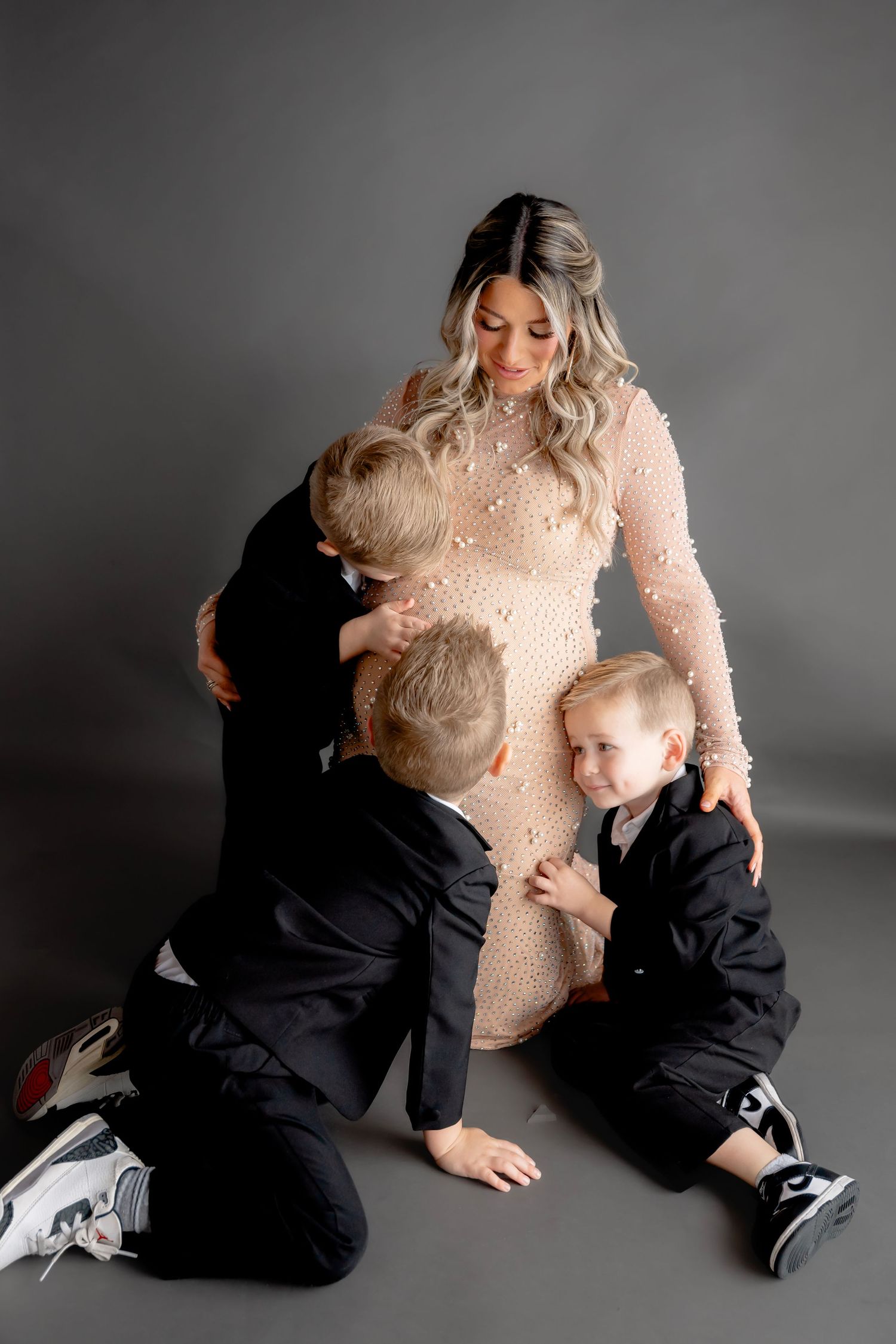 Artistic maternity photos with children in black suits hugging their mother in a beige pearl dress.